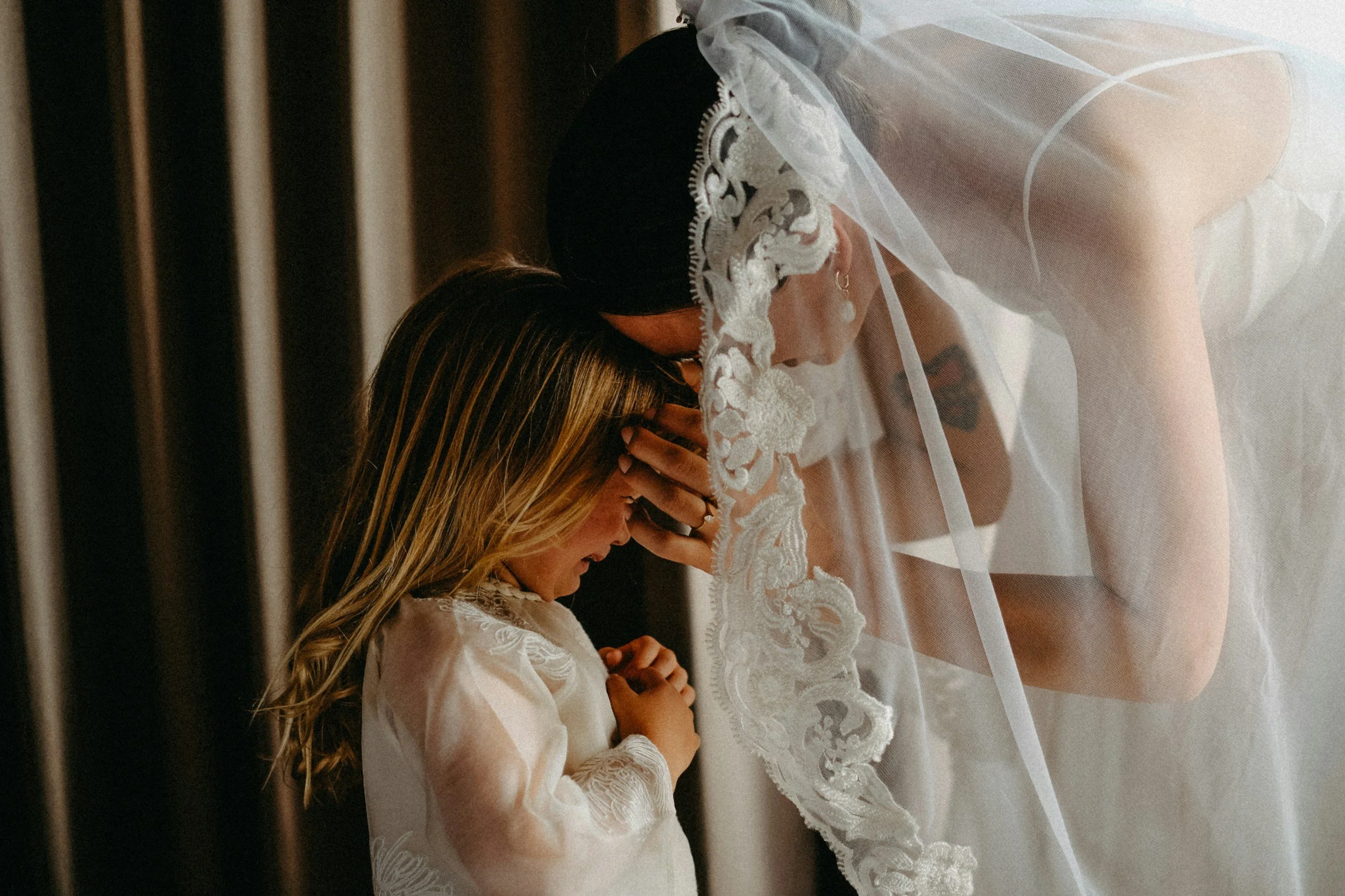 A bride and a young girl share an emotional moment, with the bride bowing her head and the girl praying, both with their hands together.