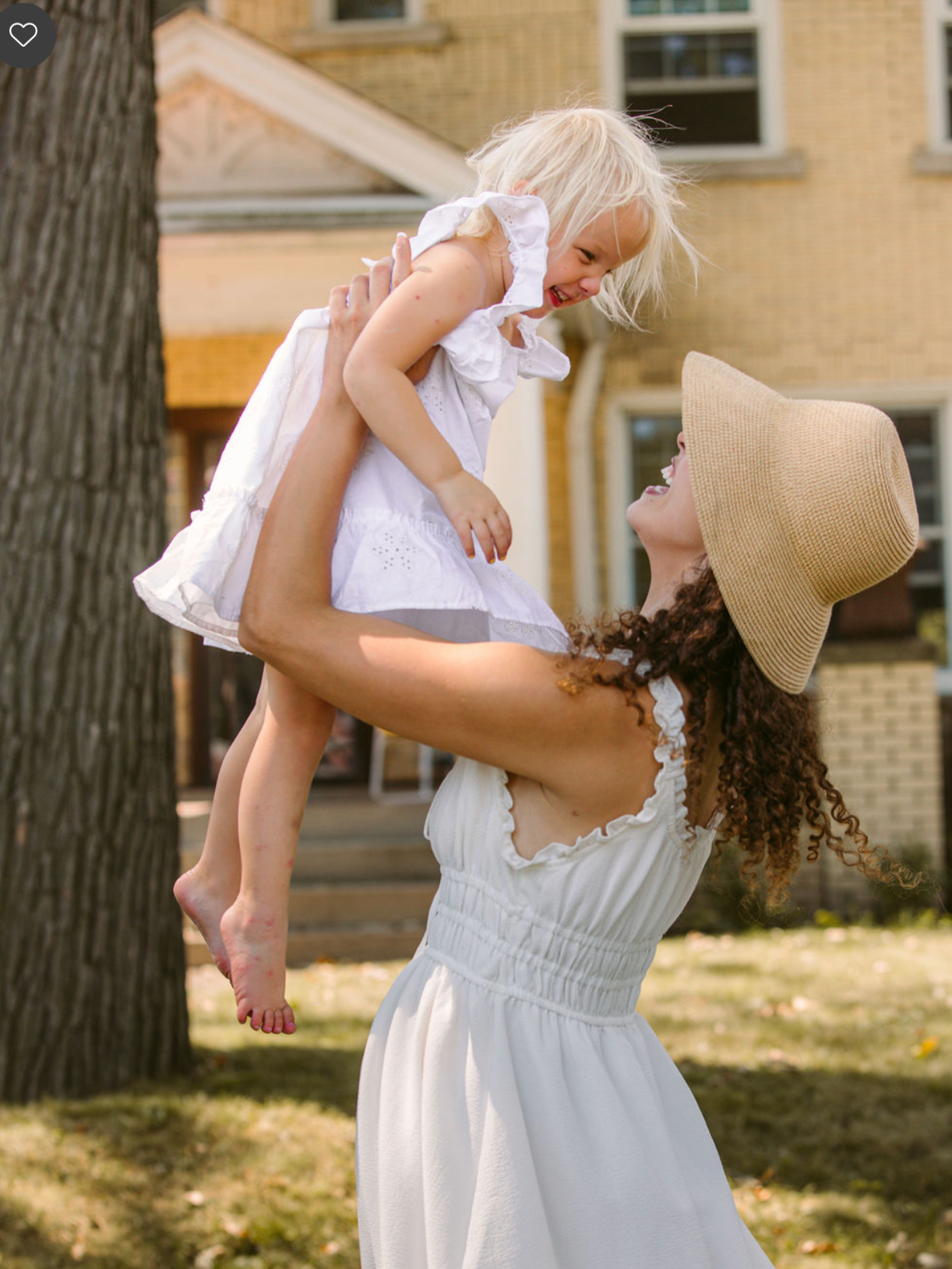Woman wearing a large sun hat and white dress lifts young girl in a white dress with ruffled sleeves. They are outdoors in a yard with grass and trees, smiling and laughing at each other.