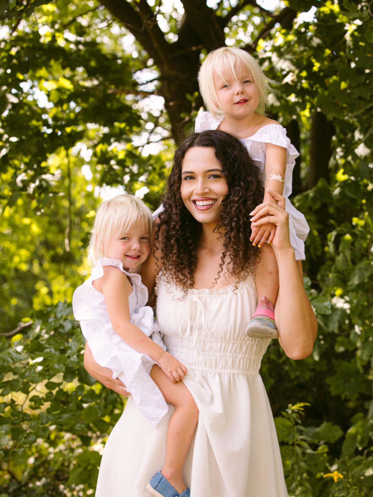 A happy woman with curly hair is smiling and carrying two young blonde girls on her shoulders outdoors near a large tree. The girls are wearing white dresses, and one girl is smiling while the other looks content. Sunlight filters through the leaves.