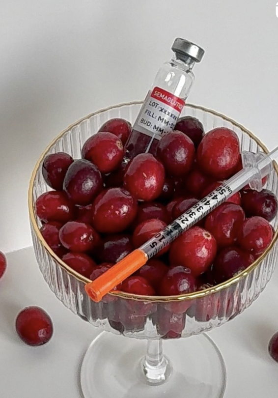 A glass bowl filled with red cherries, containing a syringe and a small vial with a label that reads "SEMAGLUTIDE."