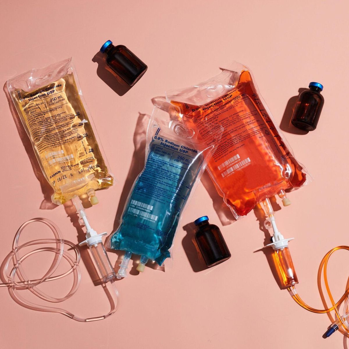Three IV fluid bags with yellow, blue, and orange liquids on a pink background, accompanied by four small brown medicine bottles and IV tubing.