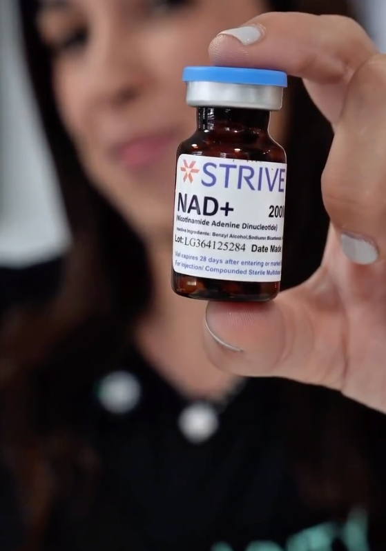 Person holding a small amber glass vial labeled 'NAD+'. The focus is on the vial, which is labeled as a dietary supplement from the brand 'STRIVE'. The person's face is blurred in the background.