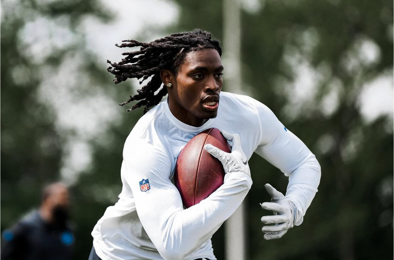 An American football player with long dreadlocks runs while clutching a football, wearing a white NFL jersey and gloves outdoors.