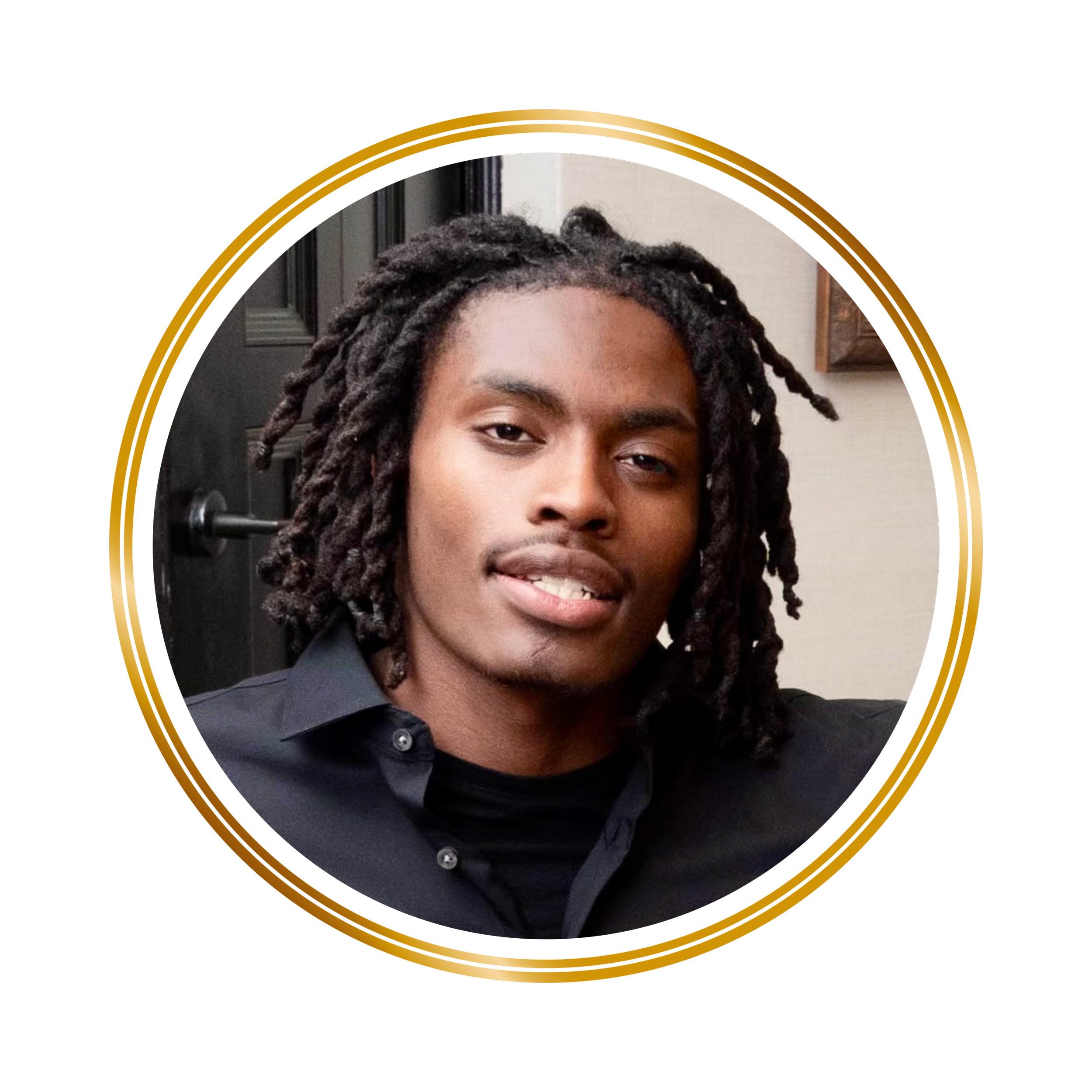 A young man with dreadlocks and a big smile, wearing a dark shirt, standing indoors with a dark door and a wall in the background.