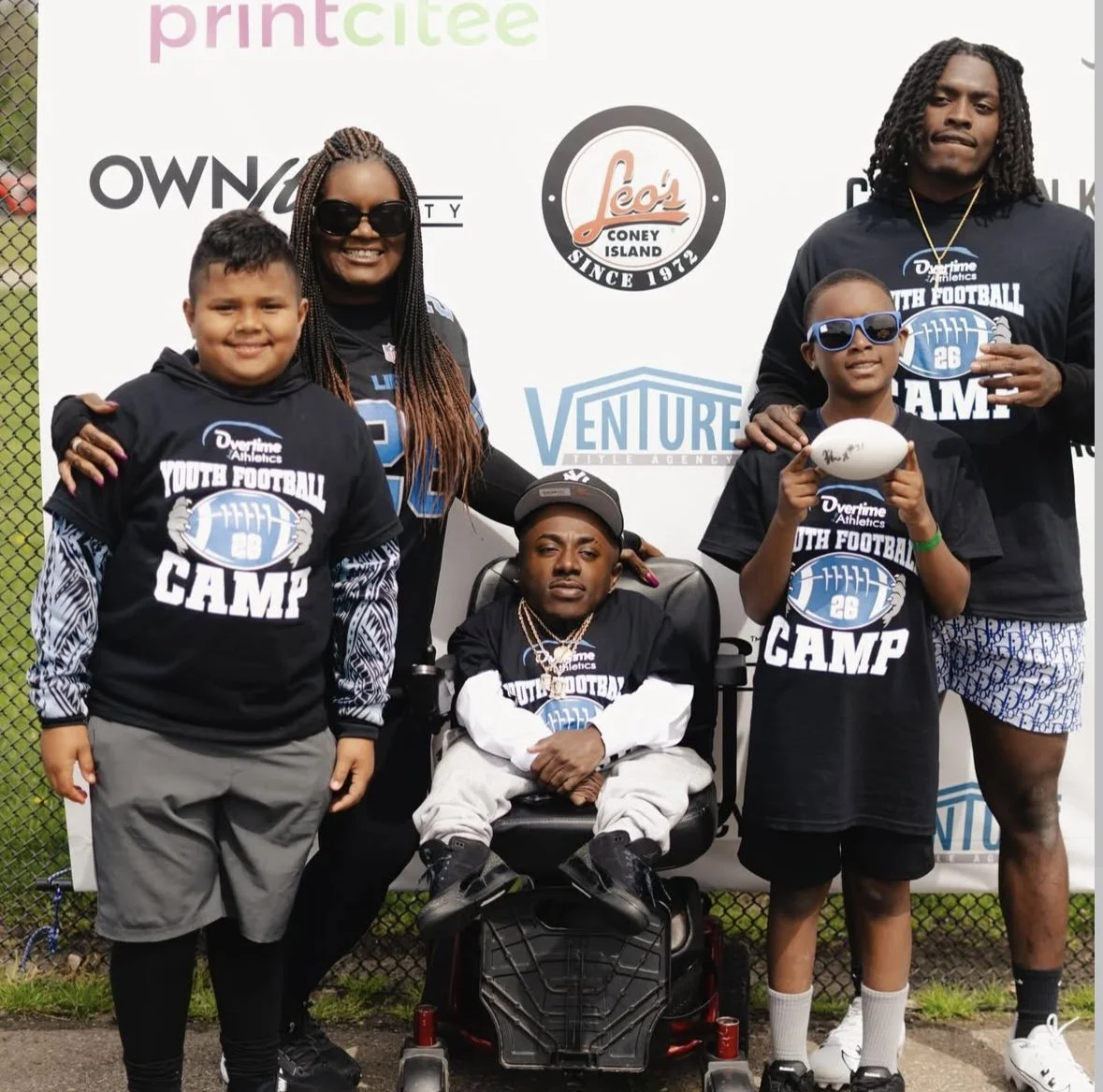 Group of football players and kids at a youth football camp, posing outdoors in front of a banner with various sponsor logos.