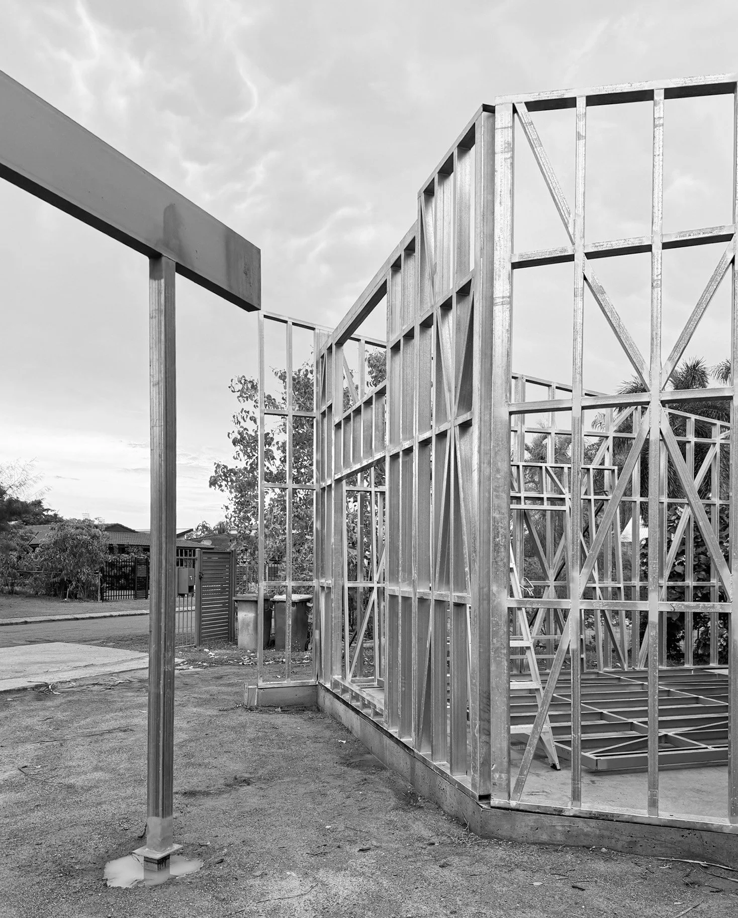 Black and white photo of a house under construction with a wooden frame. The frame is partially completed, with vertical and horizontal beams, and some diagonal supports. There is a sidewalk and street in the background, along with trees and a gated yard.
