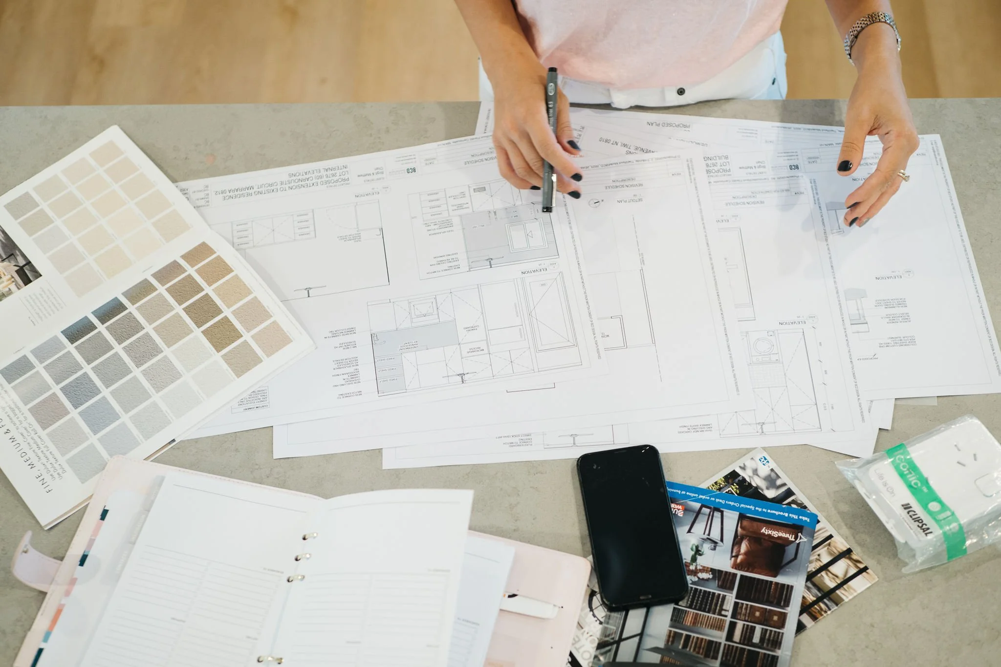 A person working on architectural plans at a desk, with color swatches, brochures, a phone, and a notepad.