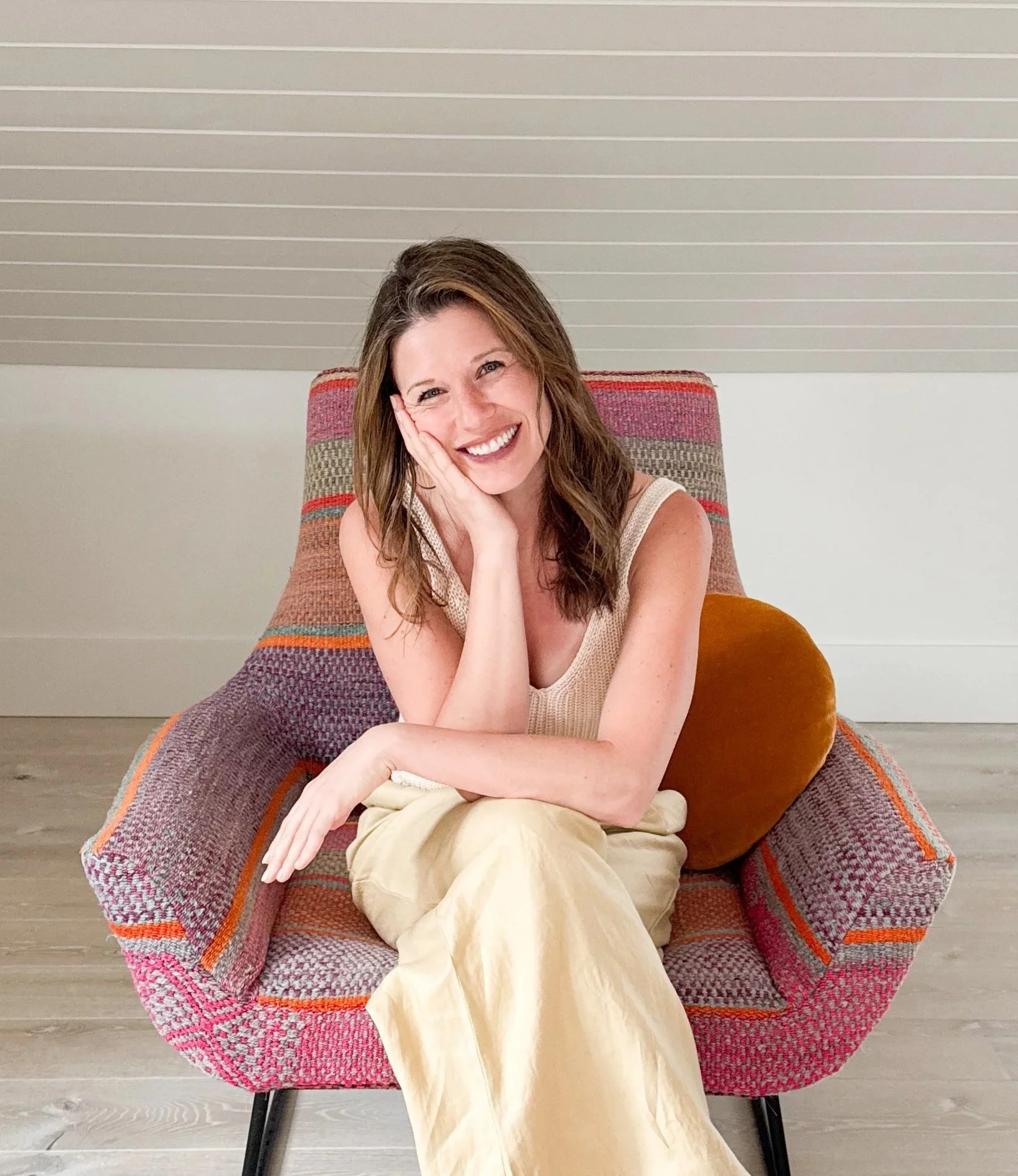 A woman with brown hair smiling, resting her head on her hand, sitting on a colorful, patterned armchair with a rust-colored pillow.