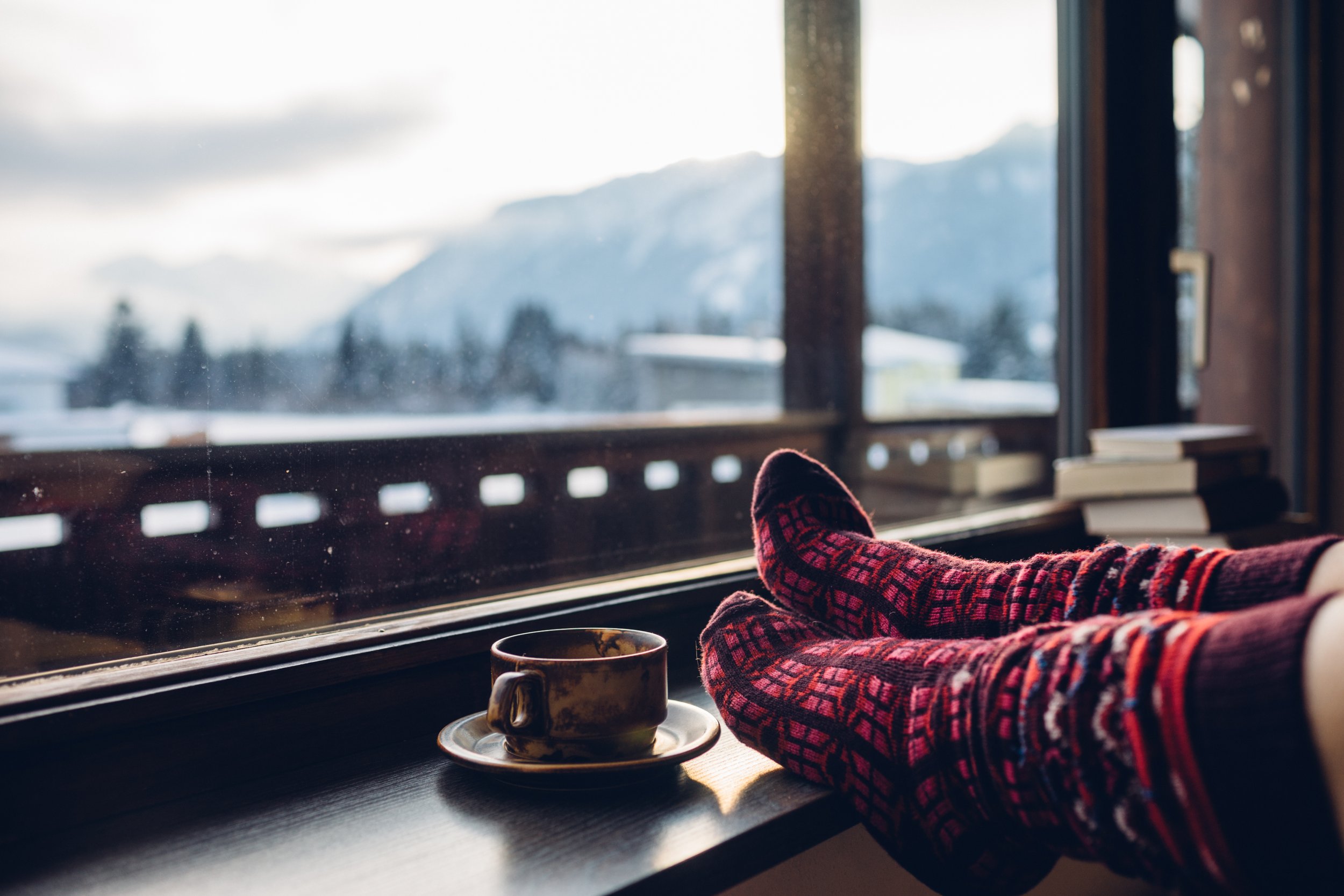 Person relaxing by a window with feet up, wearing red and black patterned socks, with a cup on the windowsill and snowy mountains outside.
