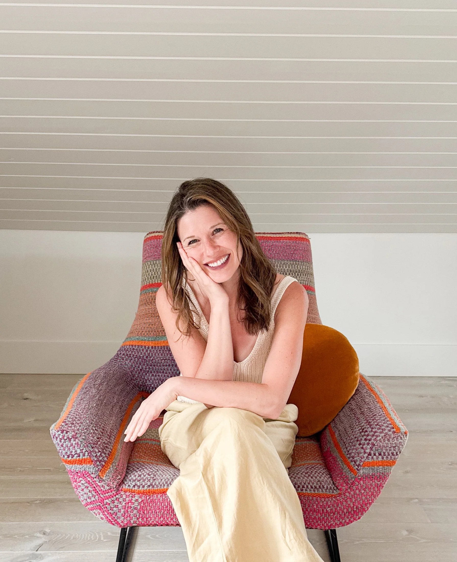 A woman sitting on a colorful armchair, smiling, with her hand on her cheek, in a room with a paneled ceiling.