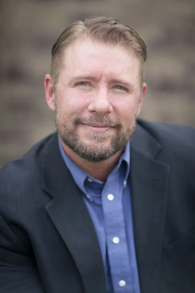 A man with light brown hair and a beard, wearing a dark suit jacket and blue collared shirt, smiling at the camera against a blurred outdoor background.