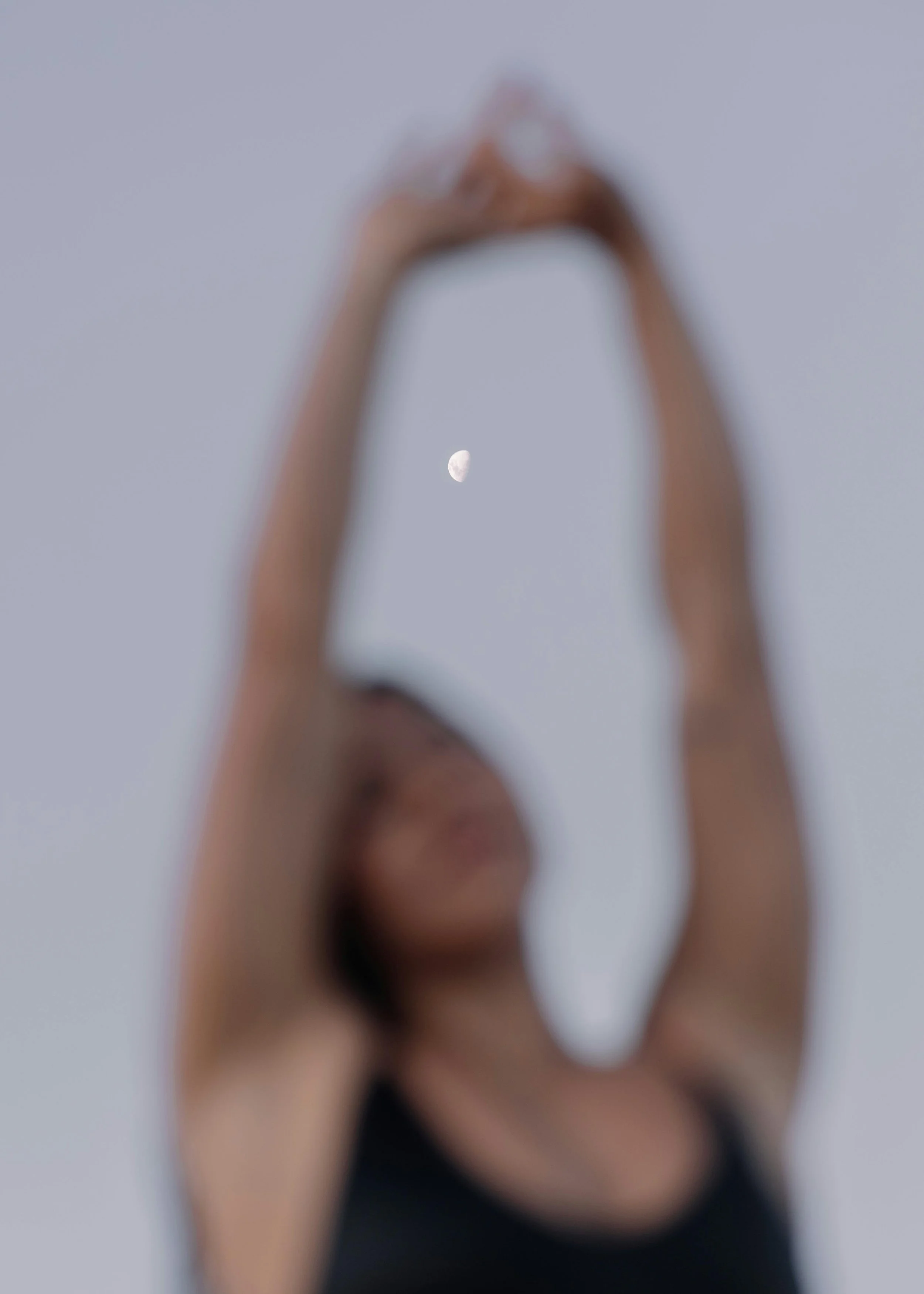 A woman with brown hair wearing a black top, holding her hands above her head, with a clear sky and a visible moon in the background.