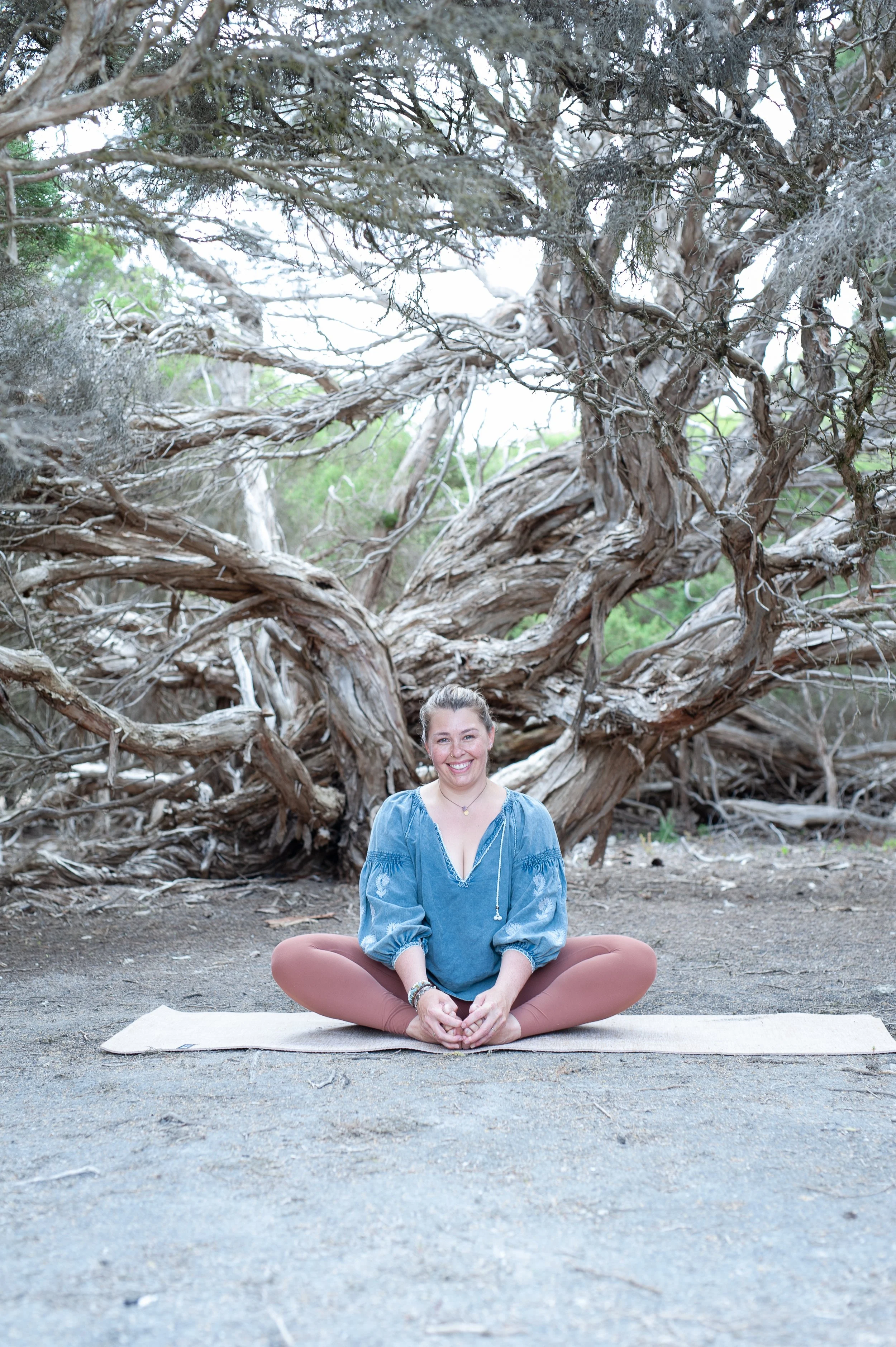 A woman sitting cross-legged on a yoga mat outdoors with a large gnarled tree in the background, smiling at the camera.