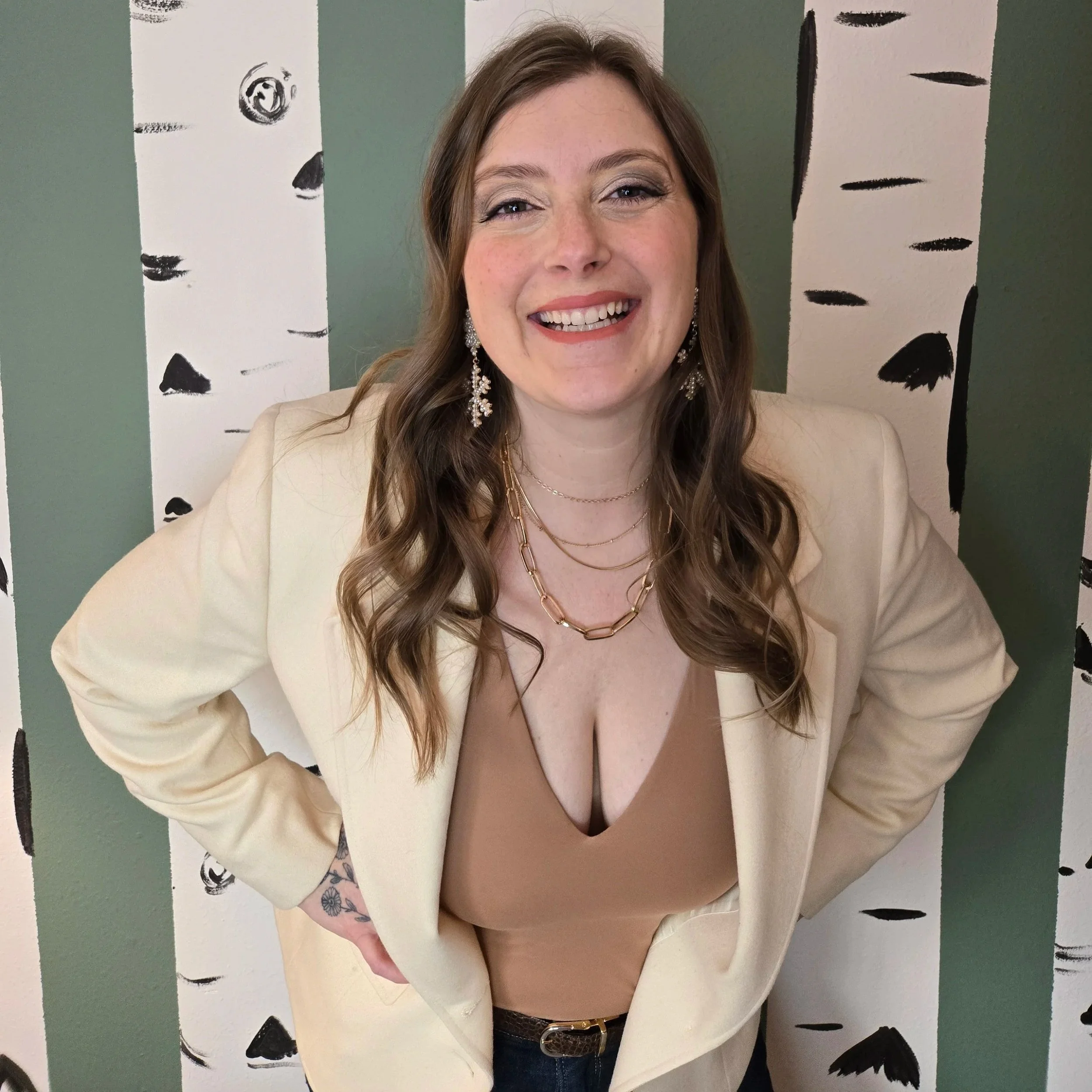 A smiling woman with light brown hair and makeup, wearing a beige blazer, layered gold necklaces, and drop earrings, standing in front of a painted wall with black and white tree bark patterns.