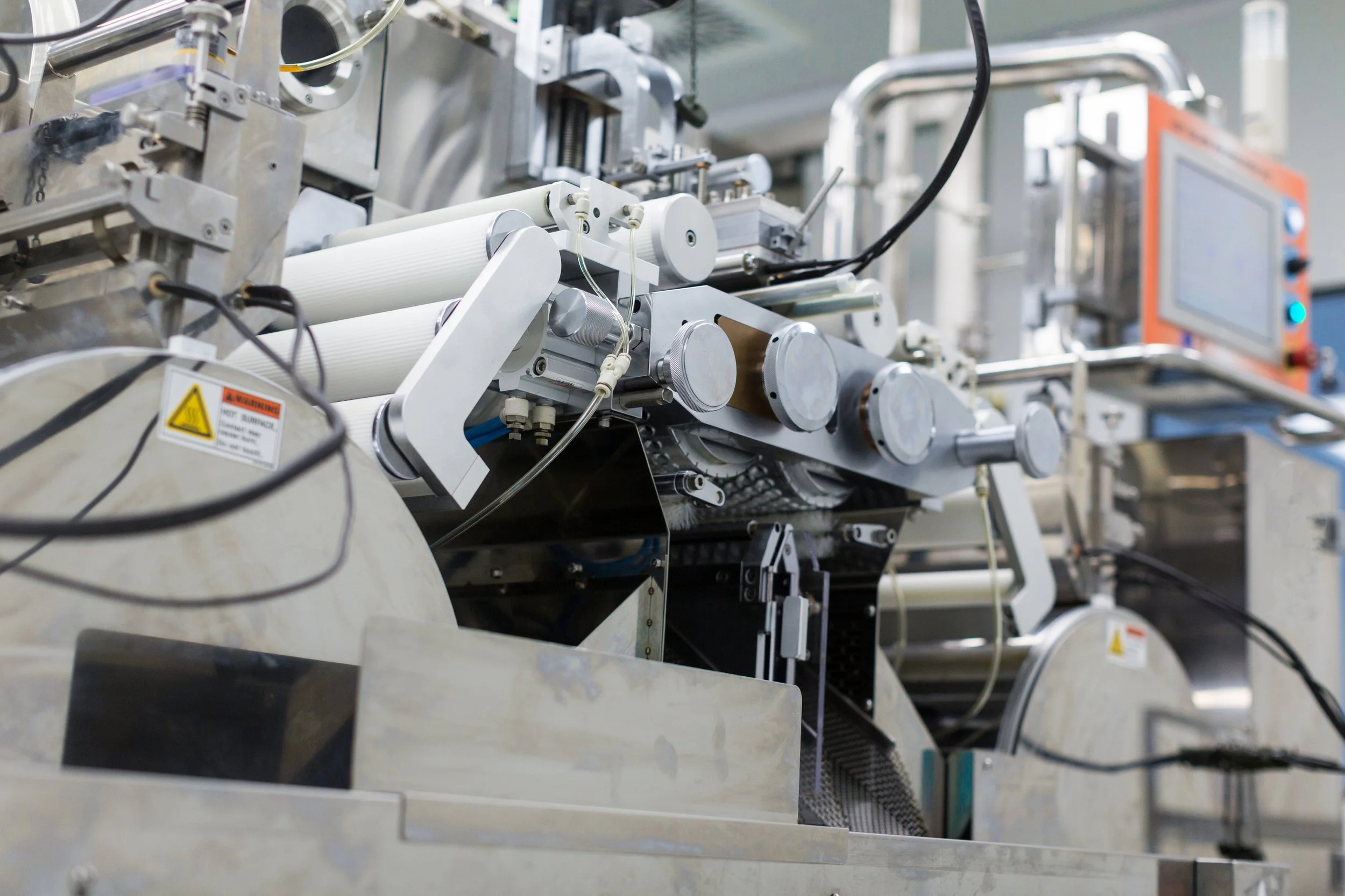 Close-up of a piece of industrial machinery, likely used in manufacturing, with various rollers, pipes, and a digital display in the background.