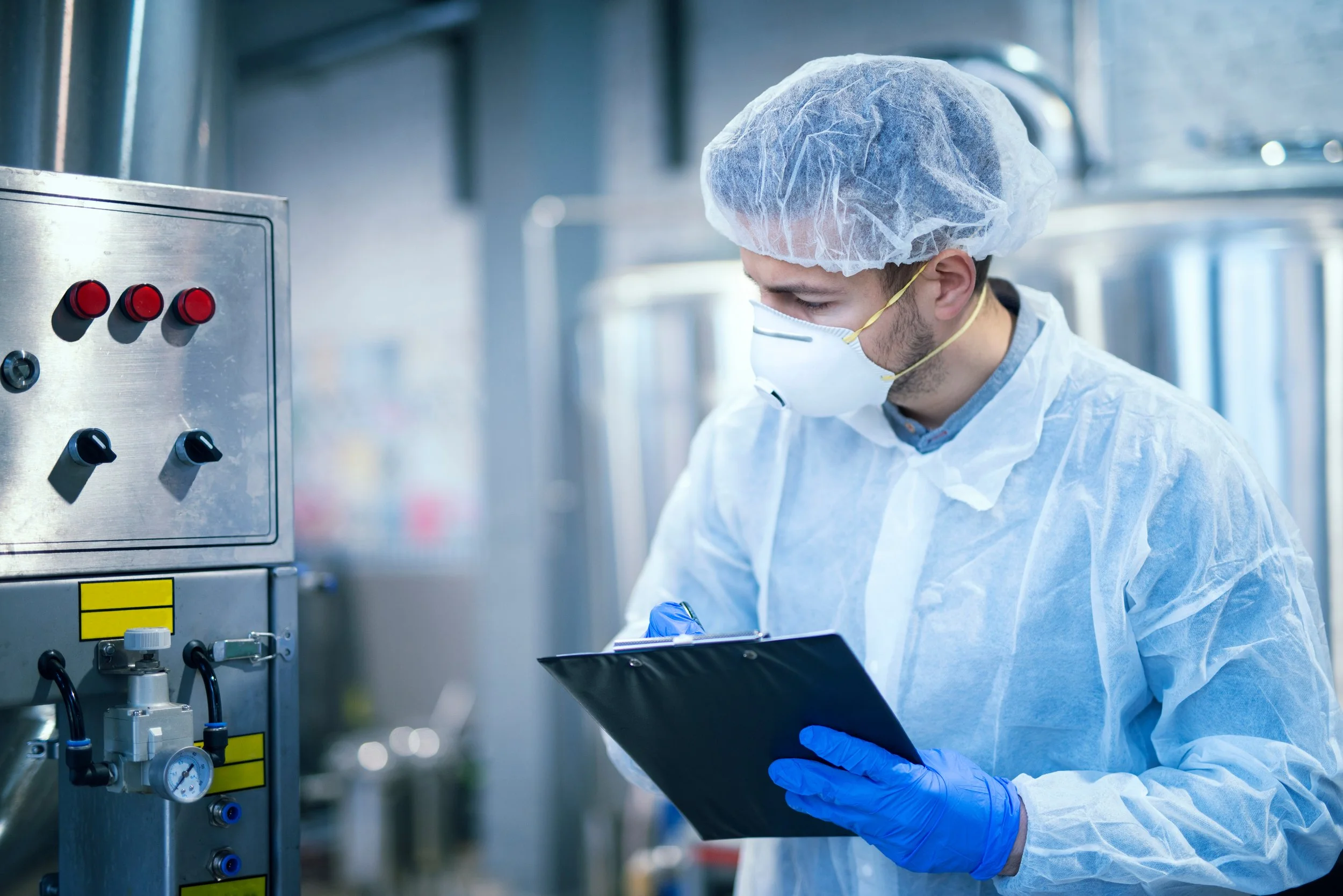 A scientist in protective gear, including a face mask, hairnet, gloves, and a lab coat, working in a laboratory, writing on a clipboard.