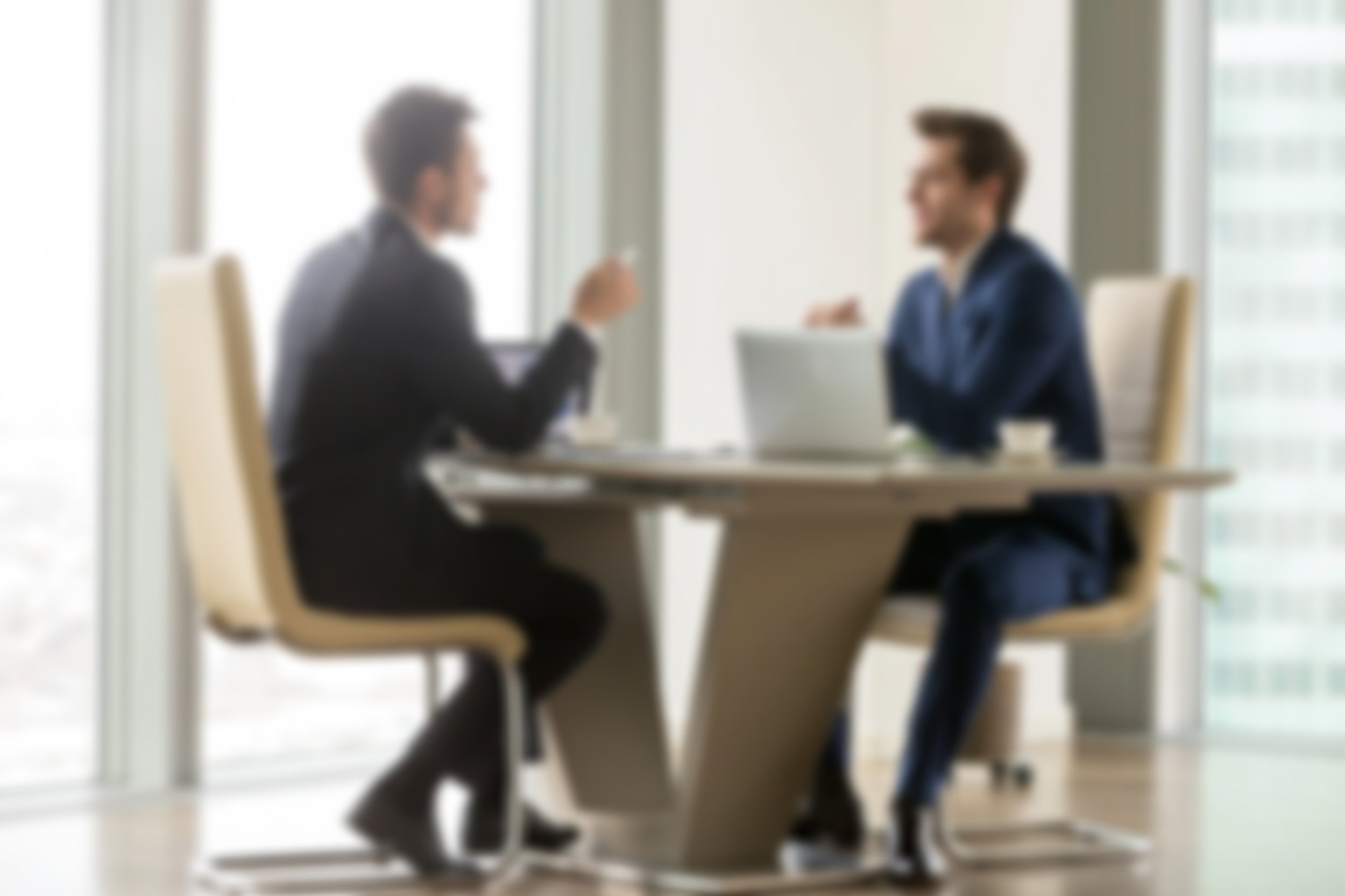 Two men in business attire having a discussion at a table in an office with large windows.