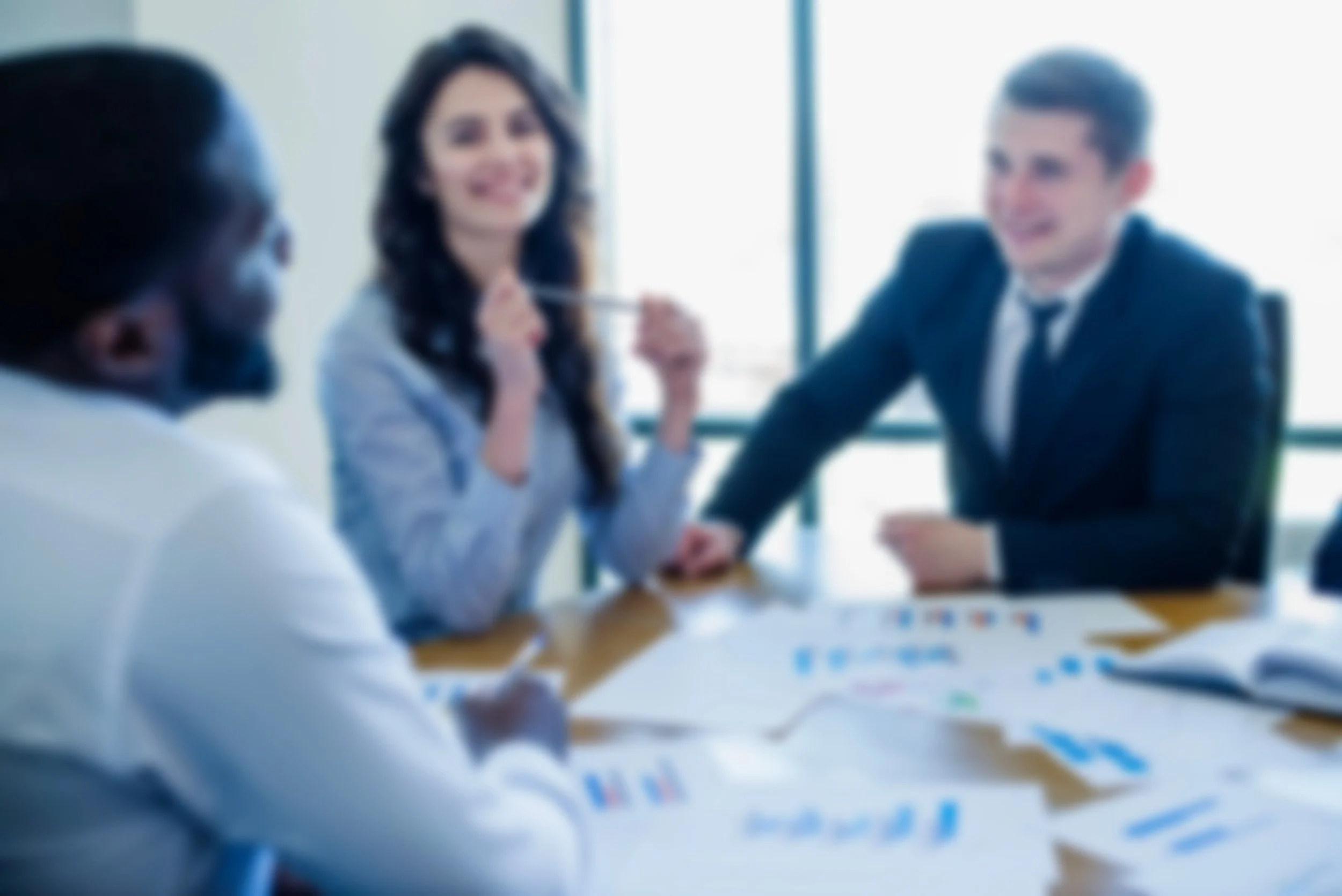 A diverse group of three business professionals having a meeting in a conference room with a large window, discussing documents and charts on the table.