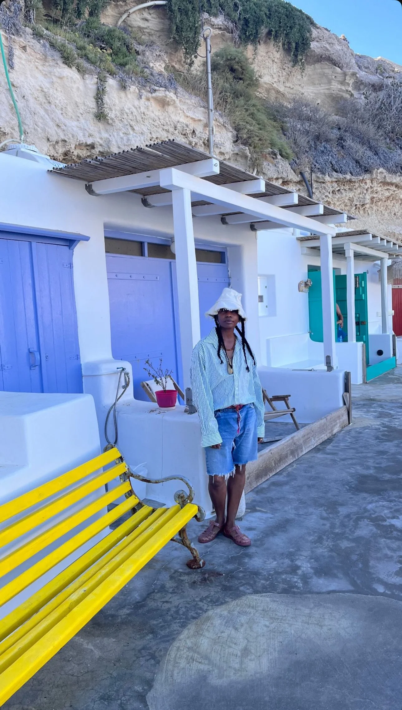A woman wearing a white bucket hat, sunglasses, a loose striped shirt, and denim shorts standing on a concrete surface with a yellow bench nearby. Behind her are white buildings with blue doors, and a rocky hillside overhead.