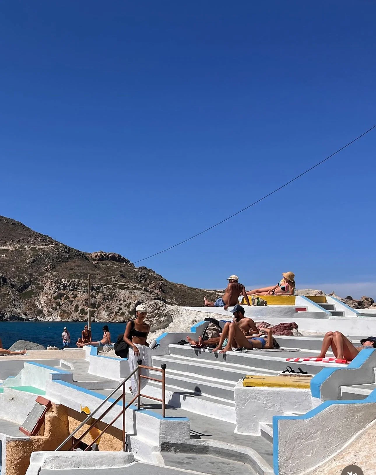 People relaxing and sunbathing on white steps with blue accents near the water on a sunny day, with a mountain in the background and a clear blue sky.