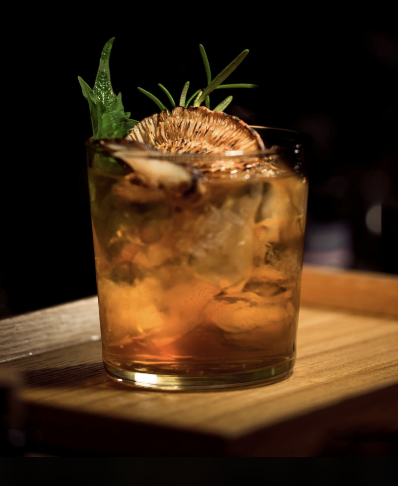 A glass filled with a whiskey-based cocktail, ice cubes, garnished with a dried orange slice and a sprig of rosemary, on a wooden surface.