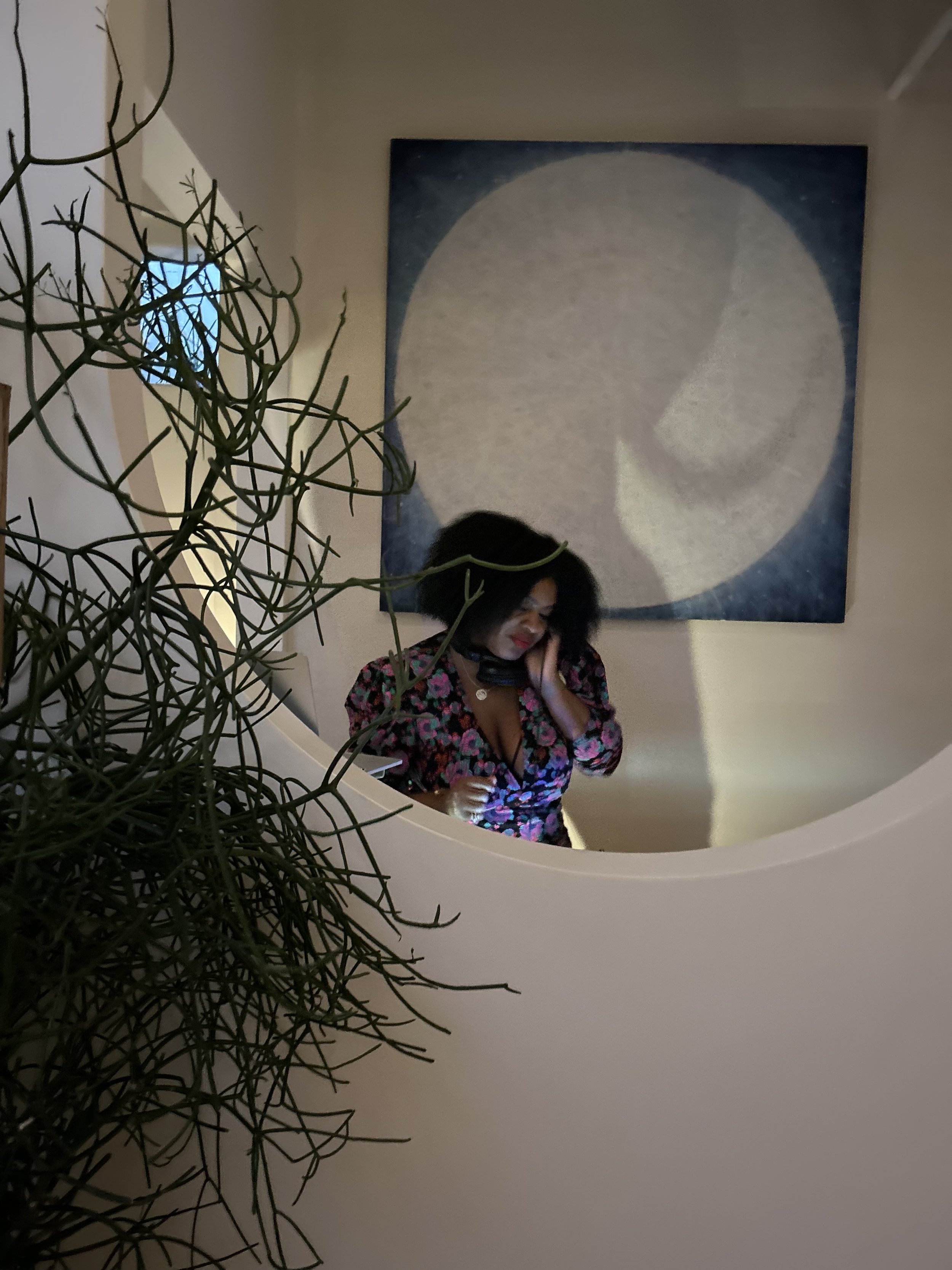 A woman with curly black hair in a floral dress, talking on the phone, is reflected in a mirror with a round frame, in a room with a large moon painting on the wall and a plant in the foreground.