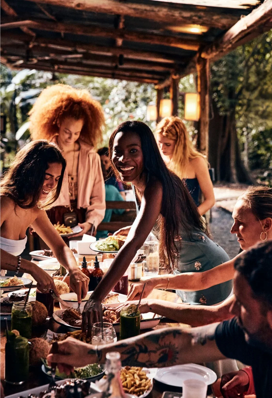 Group of women enjoying a dinner party outdoors, serving food from a long table, under a wooden shelter, with trees and hanging lights in the background enjoy Babel beverages