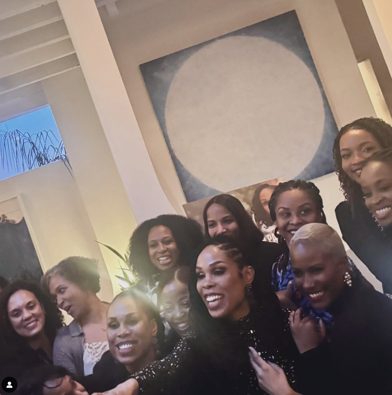 A group of women taking a selfie in a modern indoor setting with artwork on the wall and a plant in the background.
