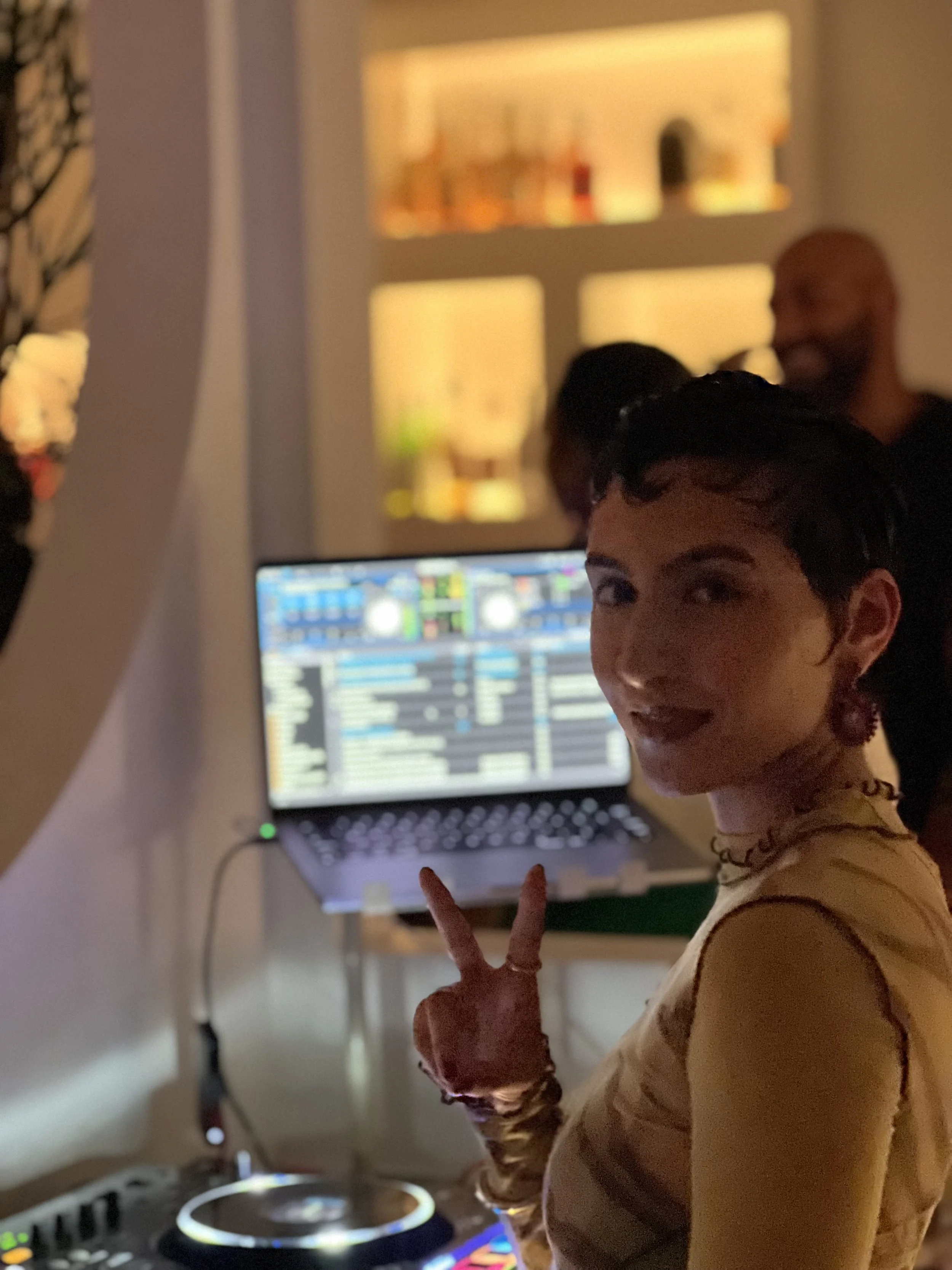 A woman with short dark hair, wearing a yellow top, making a peace sign with her right hand while smiling at the camera. She is in a dimly lit room with a DJ setup, including a laptop showing DJ software with colorful waveforms and controls, and a DJ