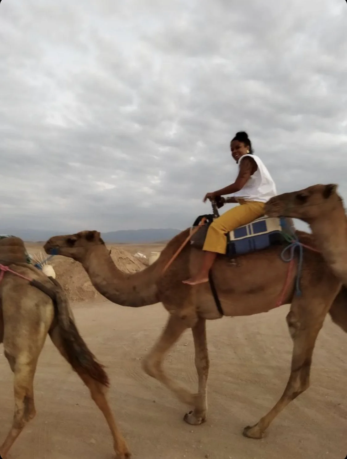 A Black woman riding a camel in a desert landscape under a cloudy sky. Marva Babel