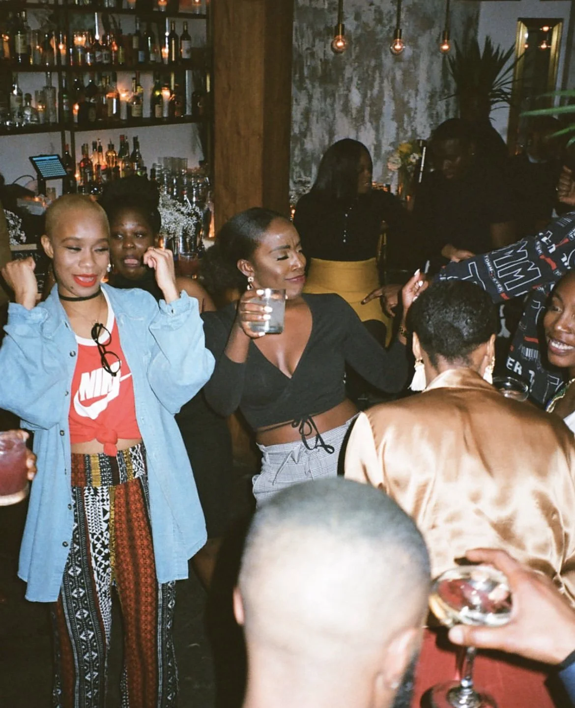 Group of people socializing and dancing at a bar or nightclub, with shelves of bottles and warm lighting in the background.