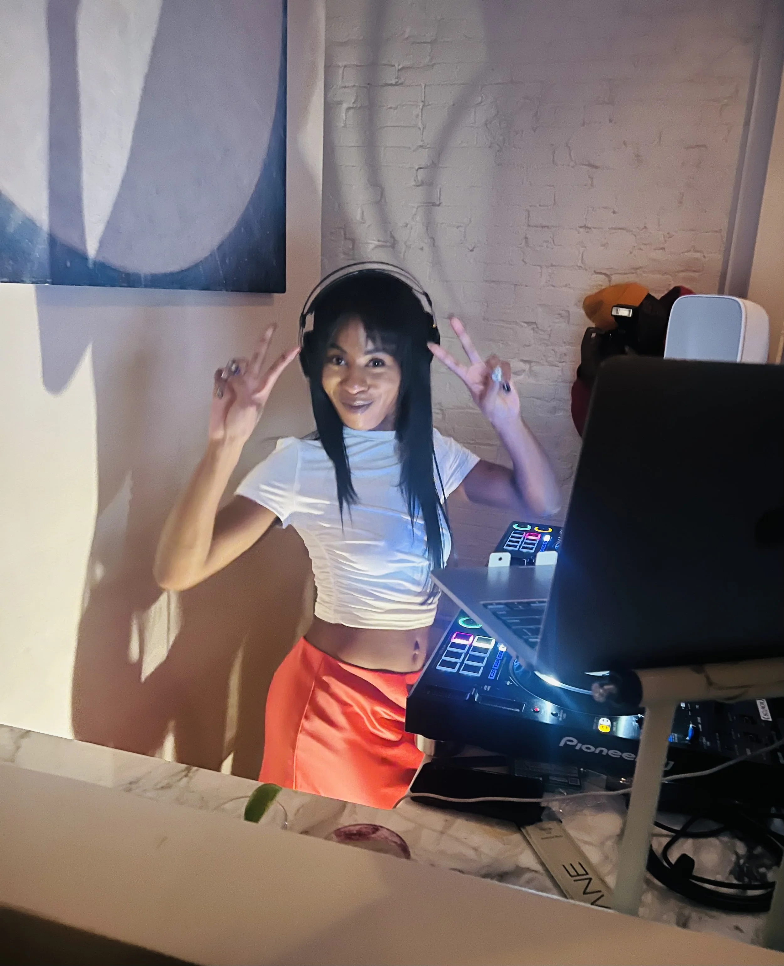 A woman with long black hair wearing a white crop top and red skirt, posing with peace signs near DJ equipment and a laptop, in a room with white brick walls and a colorful abstract painting.