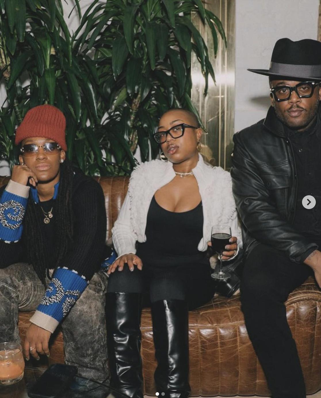 Three people sitting on a leather couch in a stylish indoor setting, with large green plants behind them. The person in the middle is holding a glass of red wine and is dressed in black with a white jacket, glasses, and jewelry. The person on the lef