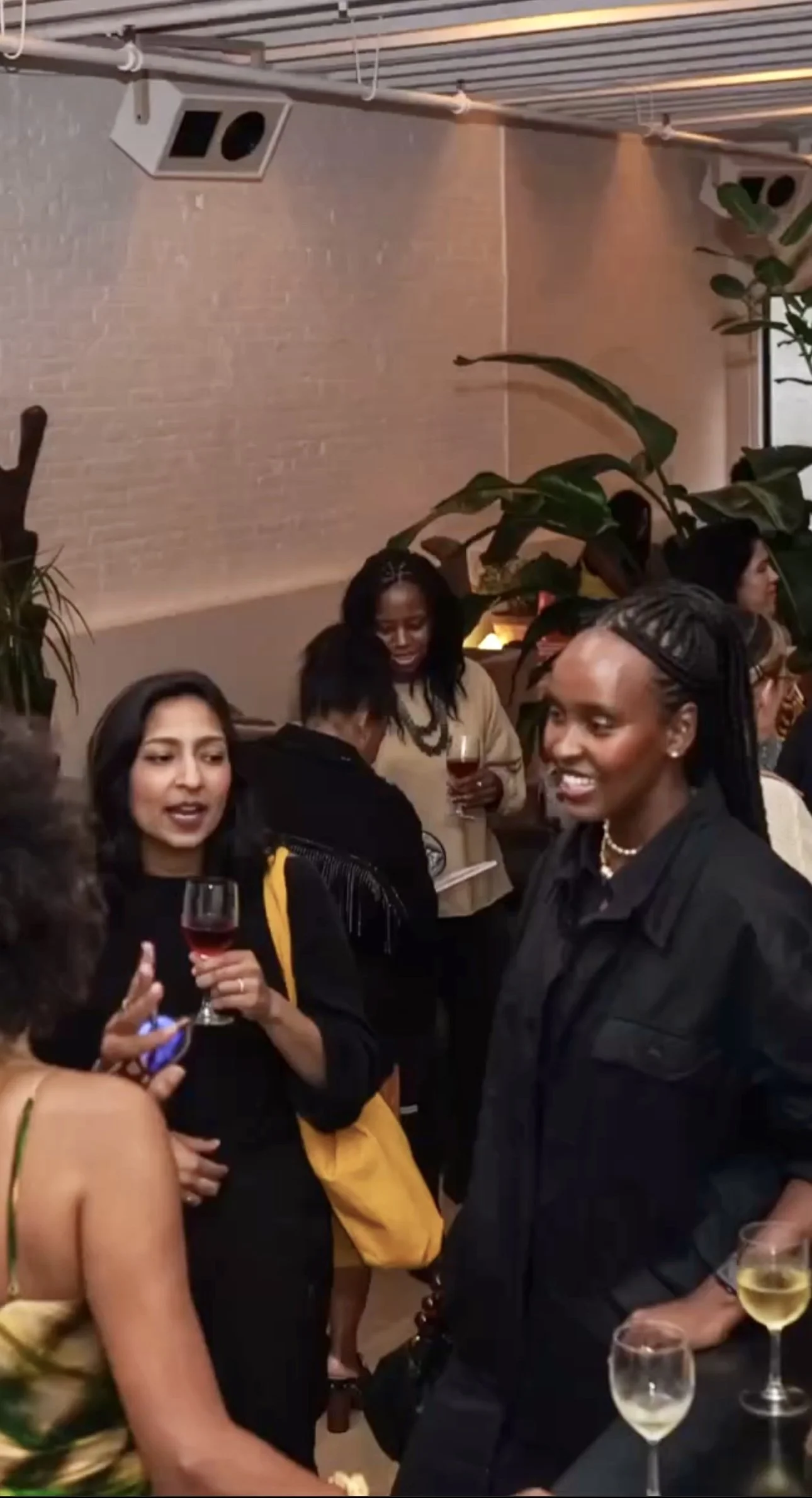 People socializing at a gathering, holding wine glasses, in an indoor venue with plants and exposed brick wall.