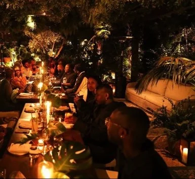 Group of people dining outdoors at night, illuminated by candlelight, surrounded by trees and plants, with a cushioned seating area in the background.