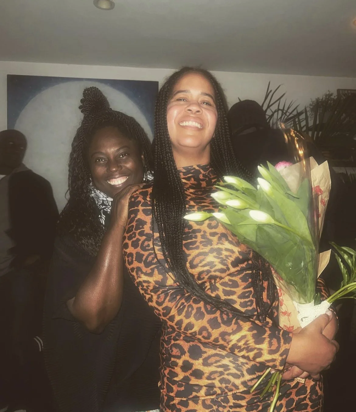 Two women smiling, one holding a bouquet of white lilies, standing together at an indoor event.