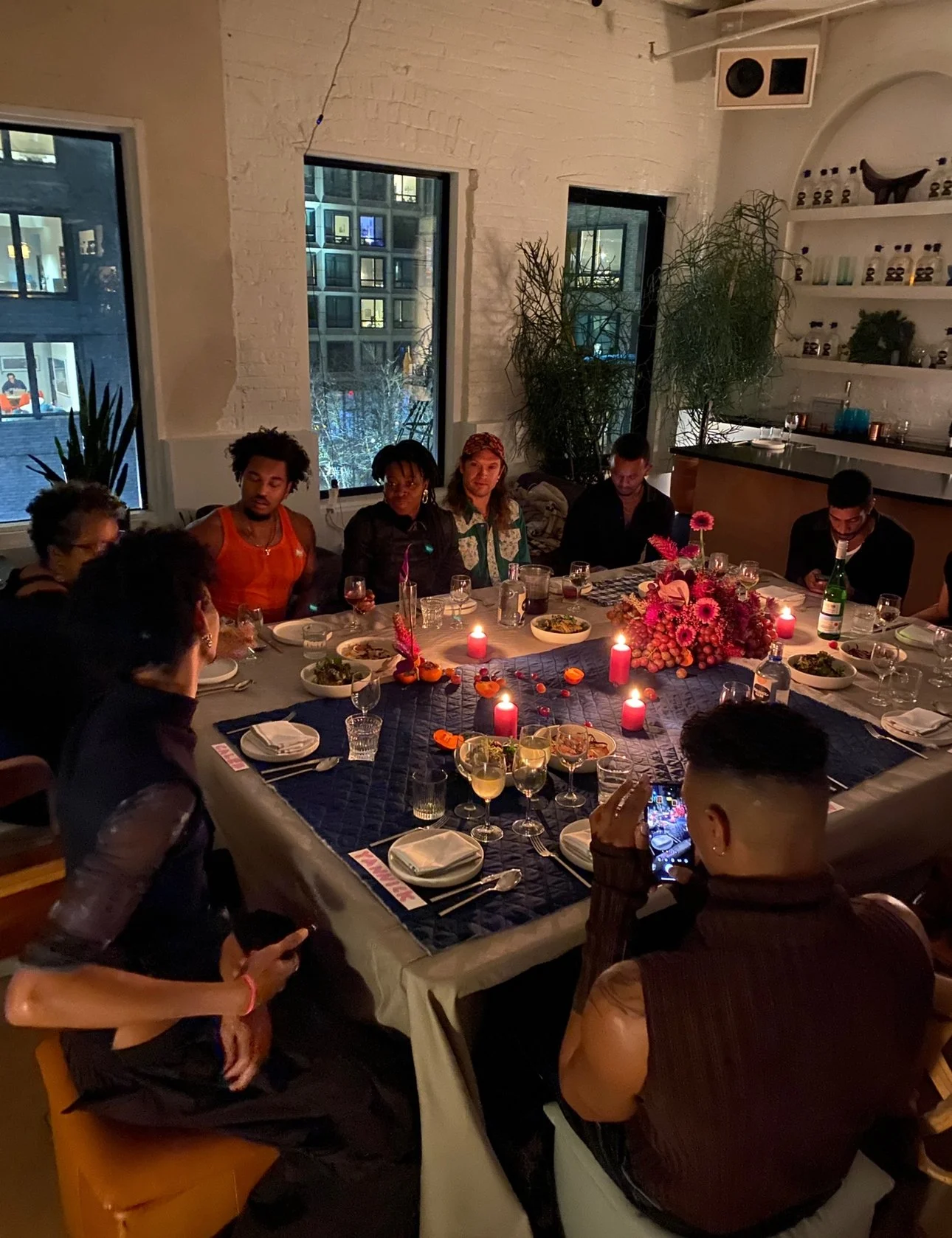 People sitting around a decorated dinner table with candles, glasses, and plates in a cozy restaurant setting with large windows and indoor plants.