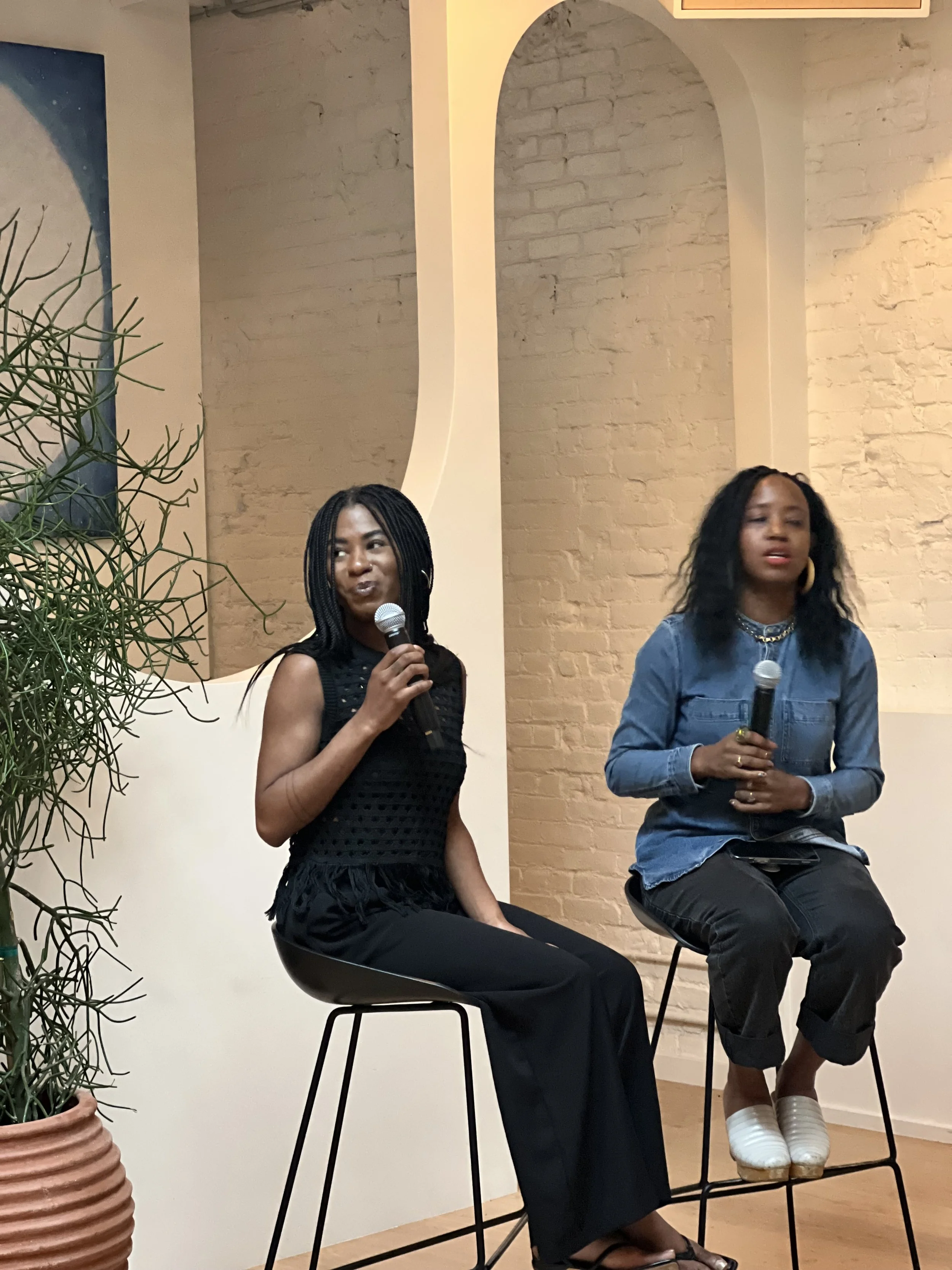 Two women of African descent sitting on bar stools holding microphones during a discussion or presentation in an indoor setting with brick walls and decorative arches.