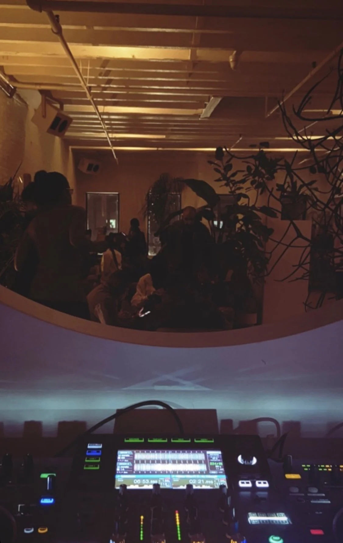 View of a dimly lit indoor space with people socializing, seen from an elevated perspective behind a DJ mixing console.