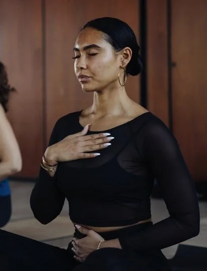 A woman with dark hair tied back, wearing a black long-sleeve top, gestures with her hand on her chest and another on her stomach, eyes closed, in a room with wood-paneled walls.