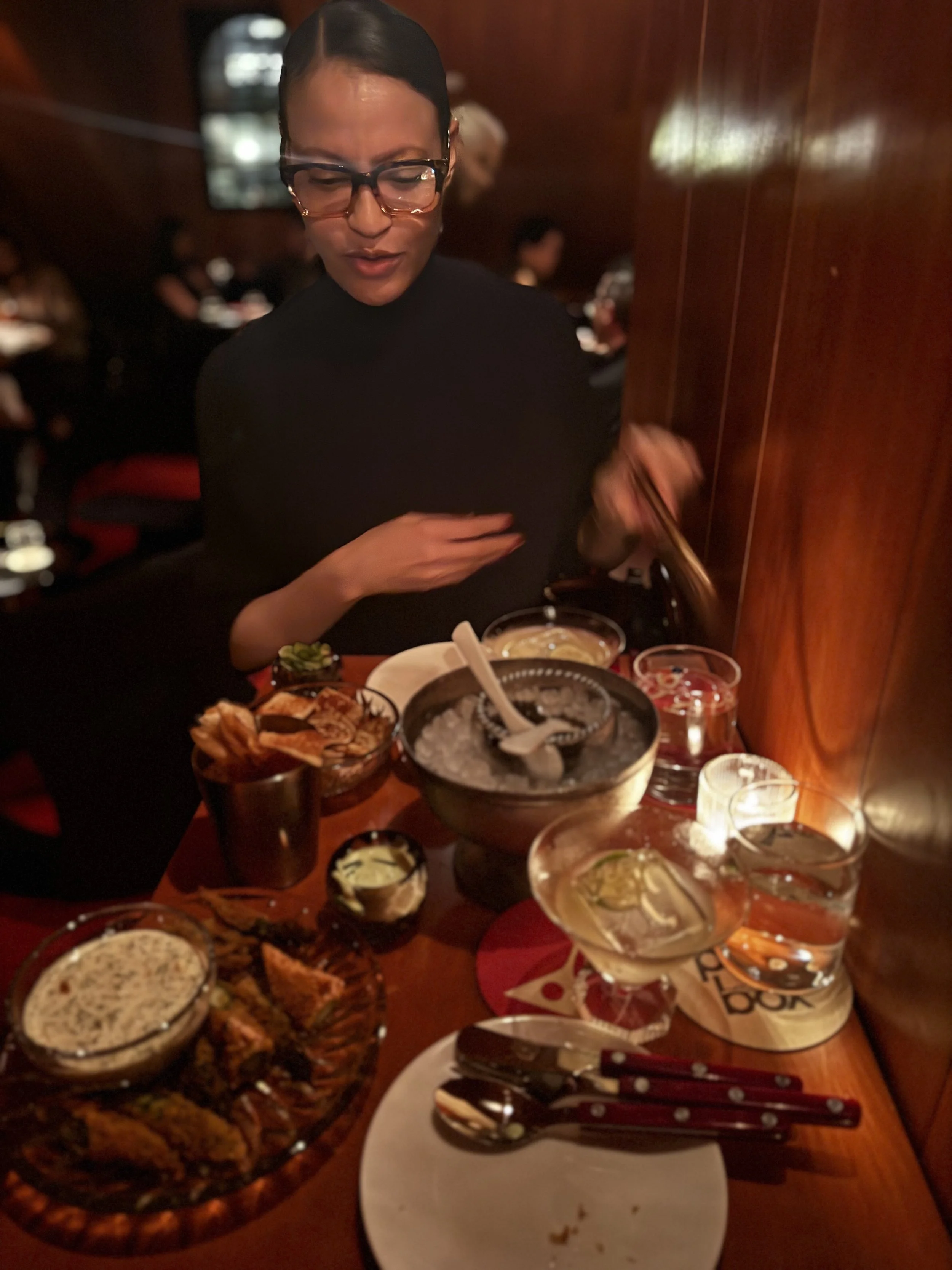 A woman wearing glasses and a black turtleneck is sitting at a restaurant table with various dishes, including a bowl of dessert, fried chicken, drinks, and condiments, in a dimly lit setting.