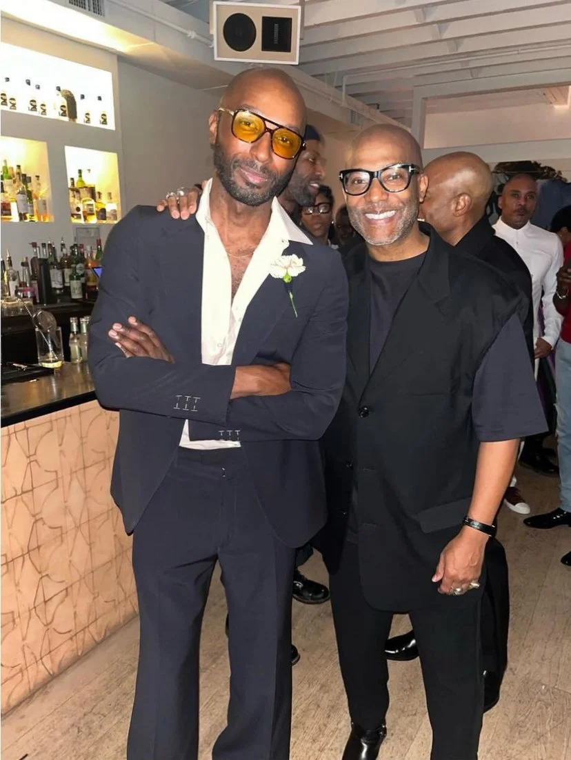Two men standing together at a social gathering, both smiling at the camera. The man on the left is wearing a dark suit with a white shirt and yellow-tinted glasses. The man on the right is wearing a black vest over a dark shirt and glasses. There ar