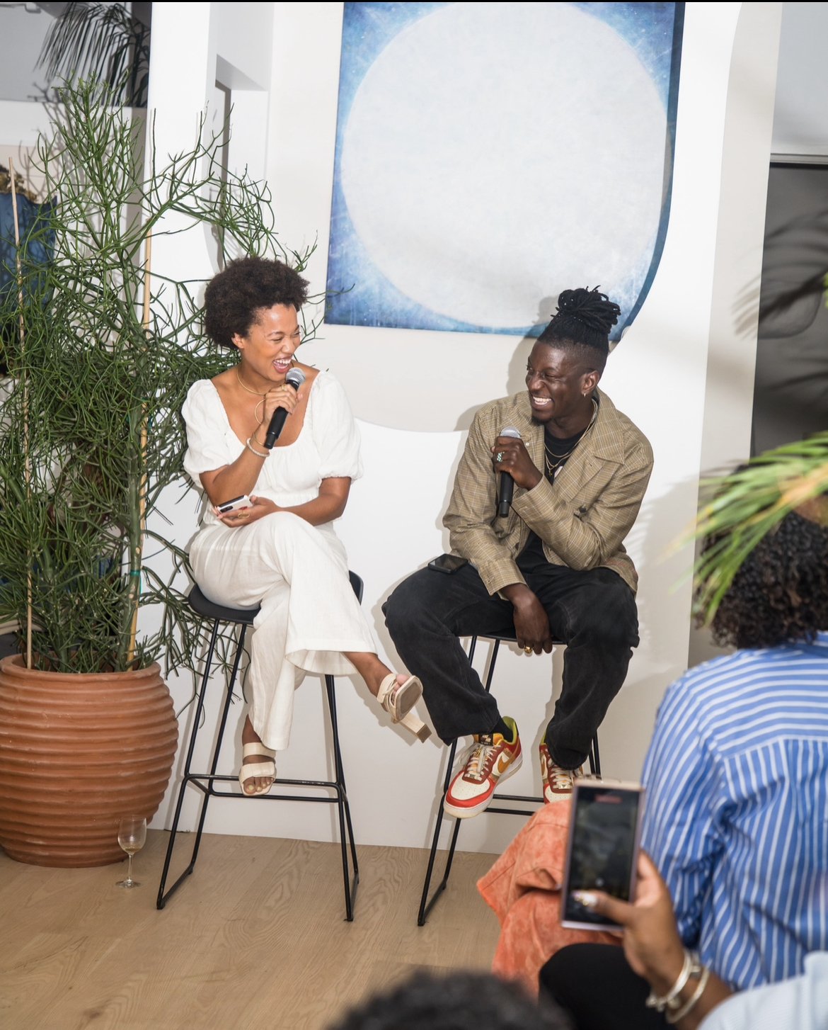 Two people sitting on high stools, laughing and holding microphones, engaged in a conversation or interview, with an audience watching and one person taking a photo, indoors with modern decor, plants, and abstract art.