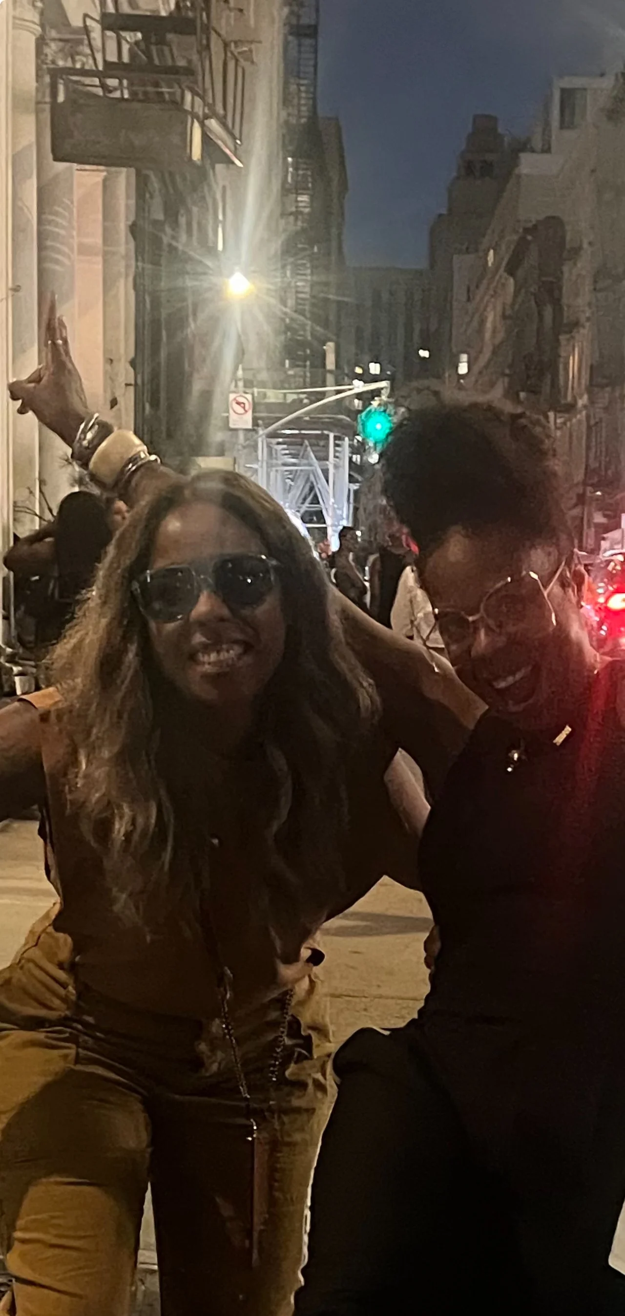 Two women smiling and posing together at night on a city street. One woman has curly hair and is wearing sunglasses, the other has short hair and is also wearing sunglasses. There are people and New York City buildings in the background.