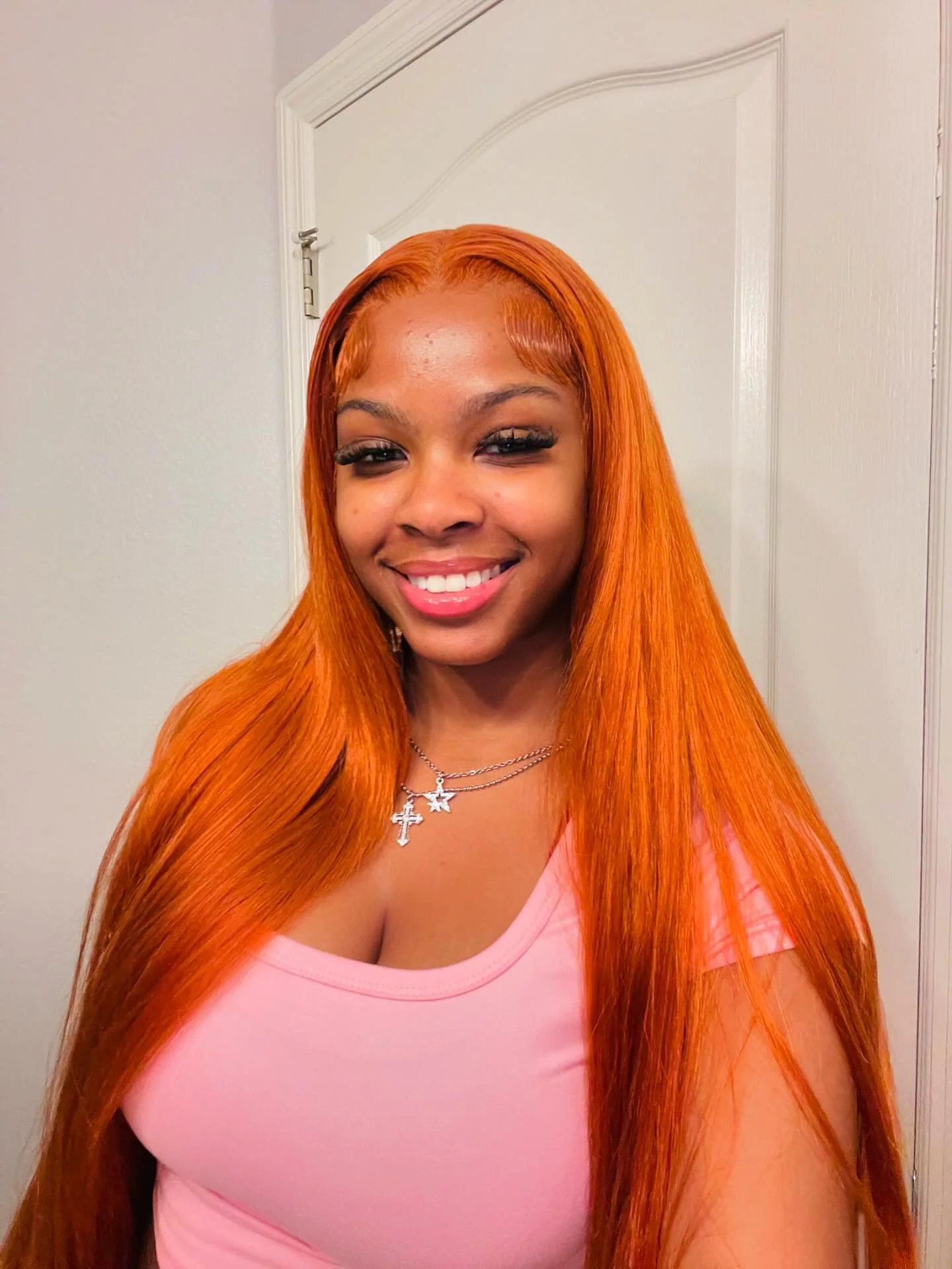 A woman with long, straight, bright orange hair smiling in front of a white door, wearing a pink top and layered necklaces with cross and star pendants.