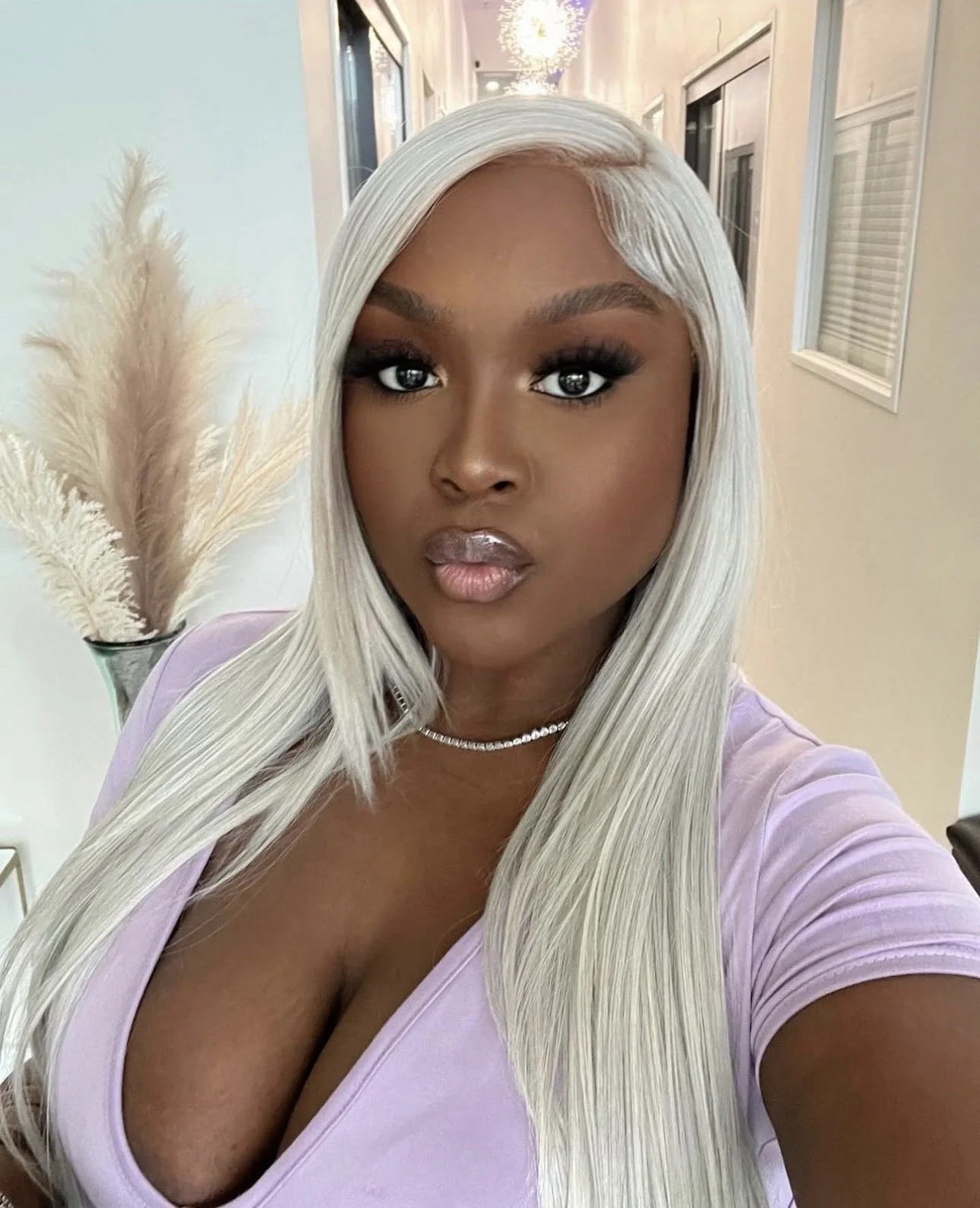 A woman with long platinum blonde hair and dark eye makeup taking a selfie in a well-lit hallway with framed pictures on the wall and a decorative chandelier hanging from the ceiling.