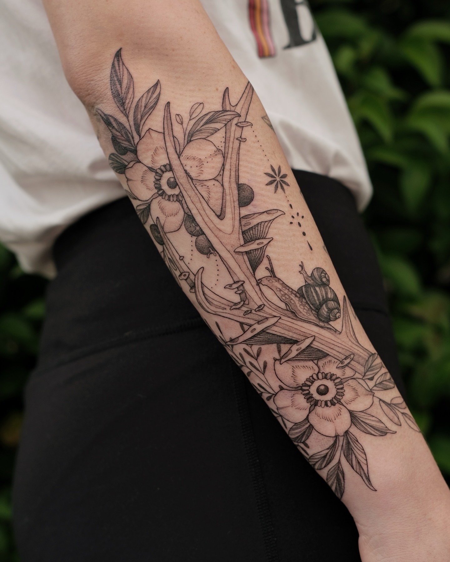 Botanical forearm tattoo featuring wildflowers, foliage, and a snail in delicate fine line blackwork.