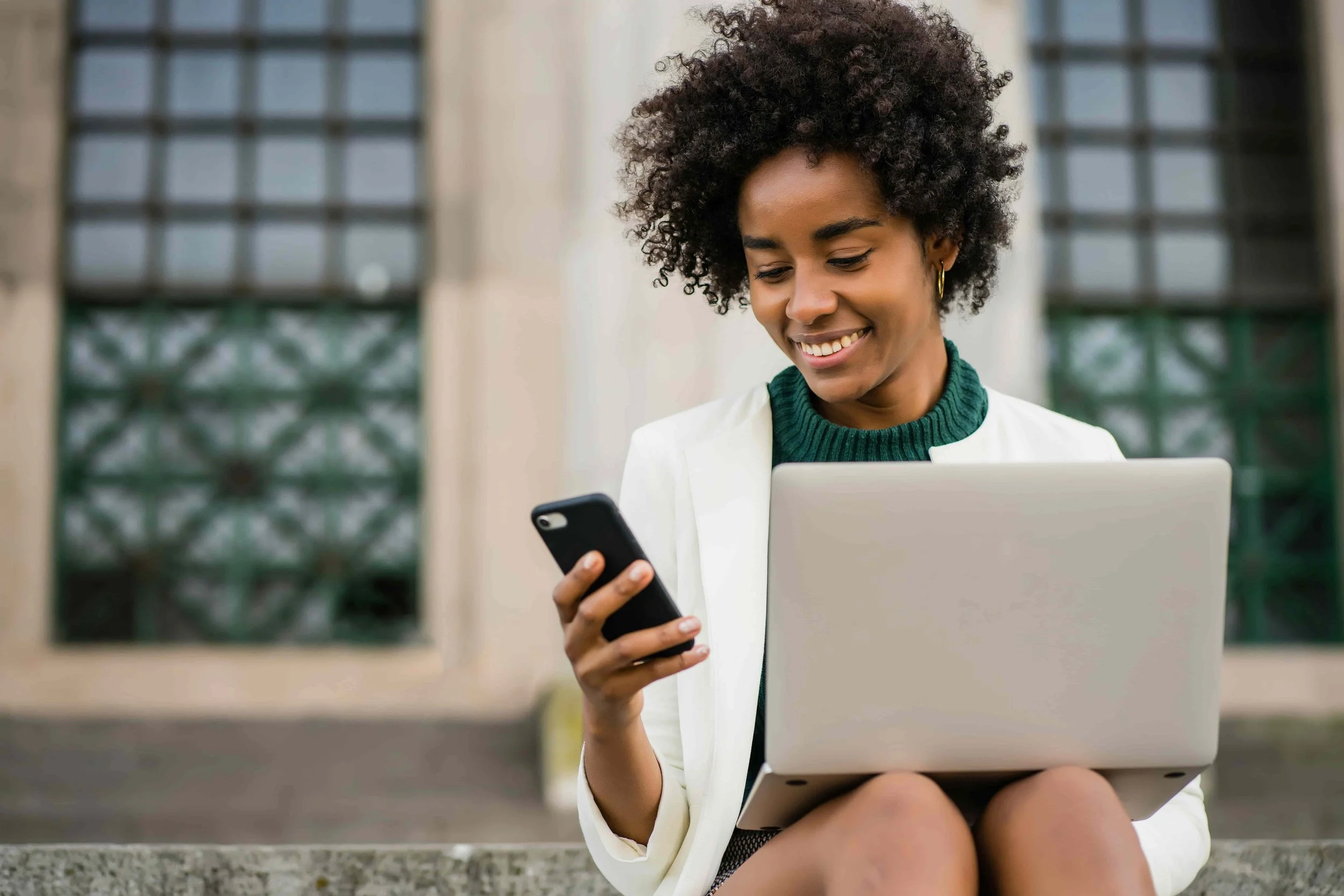 A young woman with curly hair, wearing a white blazer and green sweater, sits outdoors holding a smartphone in her right hand and a laptop on her lap, smiling as she looks at her phone.