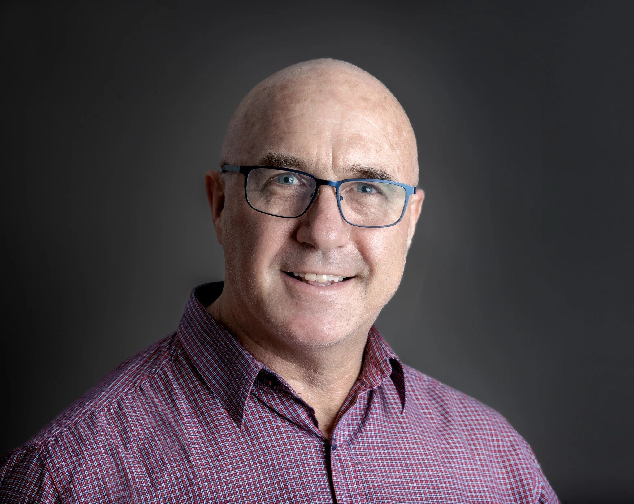 Close-up of a middle-aged man with glasses, smiling, wearing a red checkered shirt against a dark gray background.