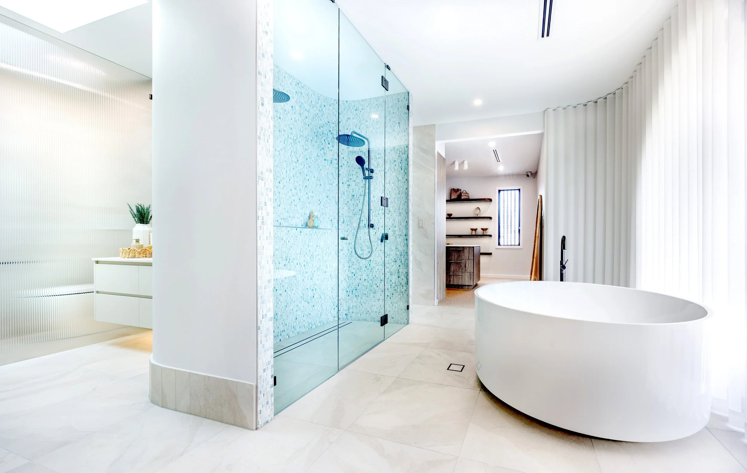 Modern bathroom with a freestanding oval bathtub, a glass-enclosed walk-in shower with blue mosaic tiles, and natural light from large windows with white curtains.
